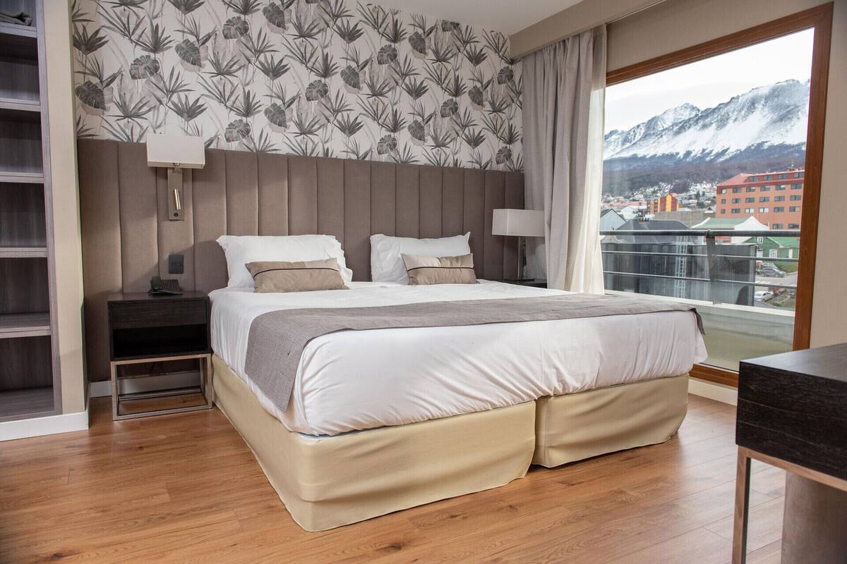 A hotel room with a bed, bedside tables, a desk, a view of mountains, and a patterned wallpaper.
