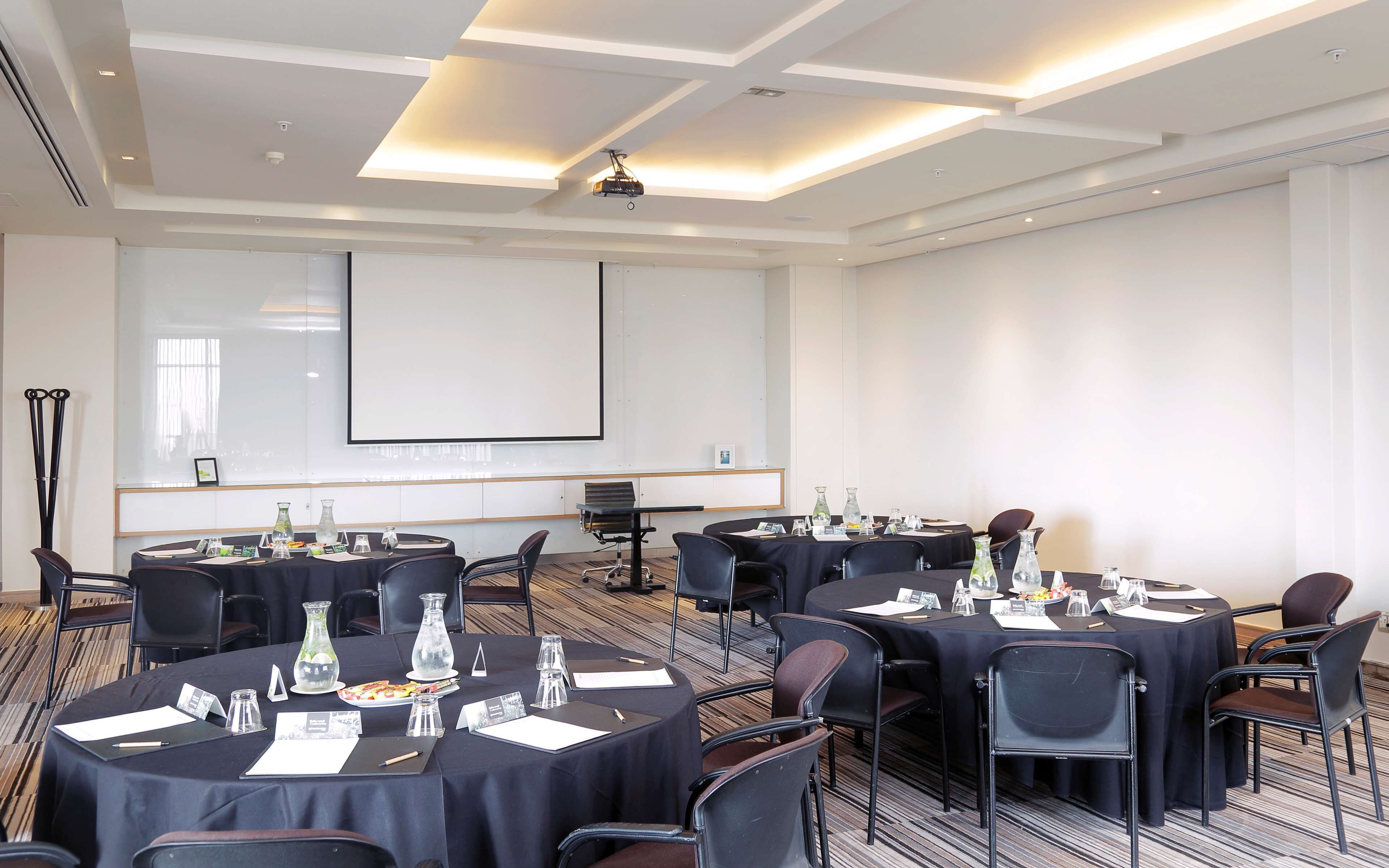A conference room with multiple tables, chairs, and a large screen for presentations.