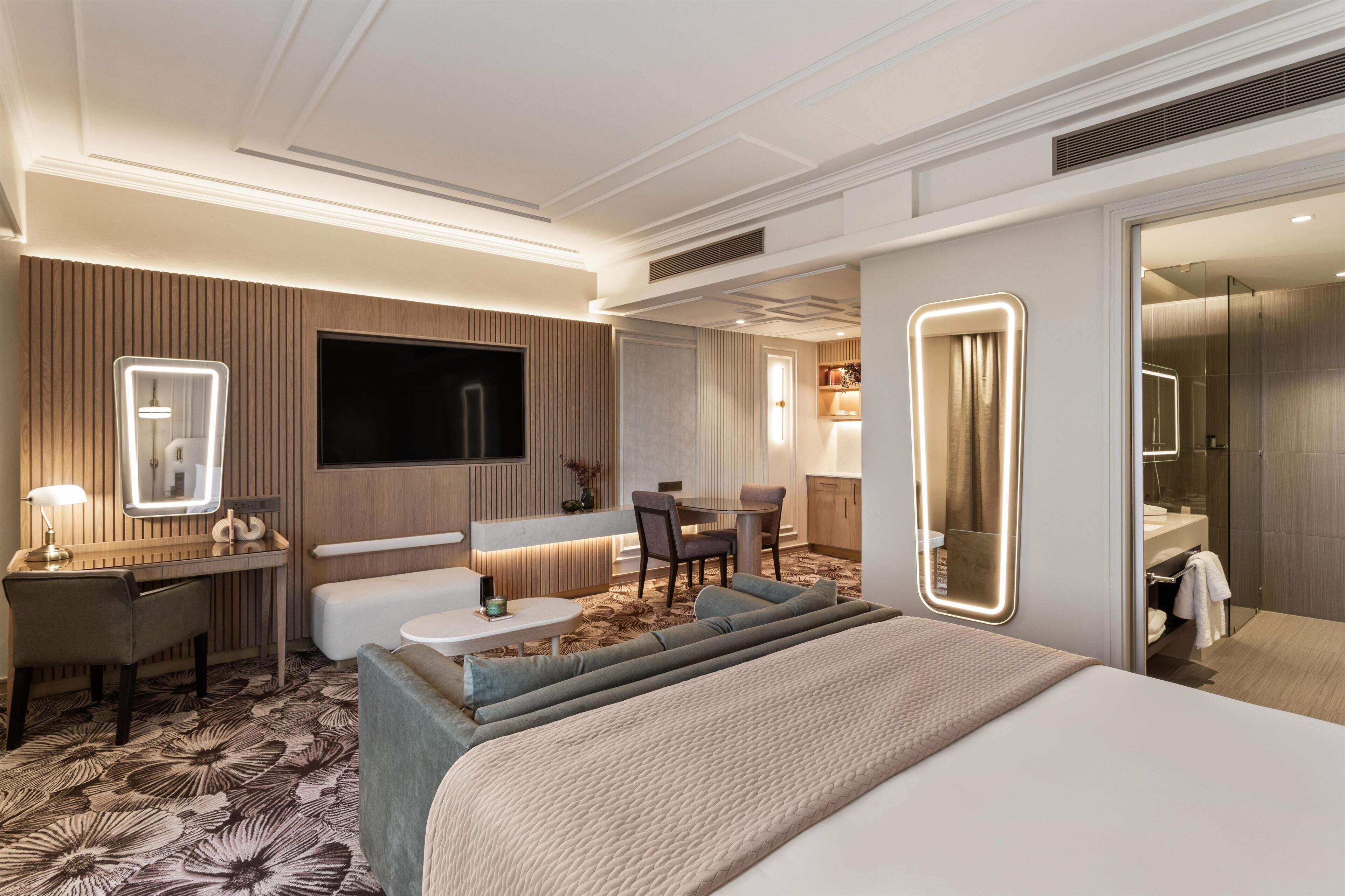 A modern hotel room with a large bed, a desk with a chair, a sofa, a TV mounted on the wall, and a bathroom visible through an open door.