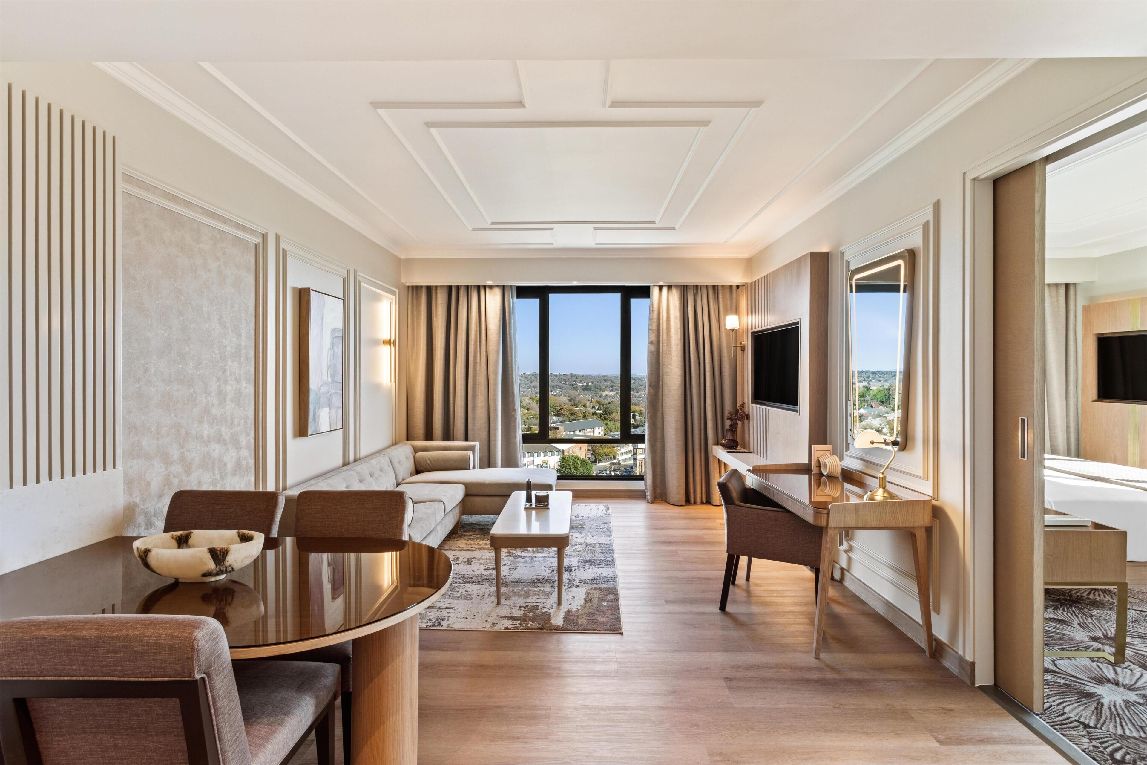 A hotel room with a sofa, a dining table, a TV, and a view of the city.