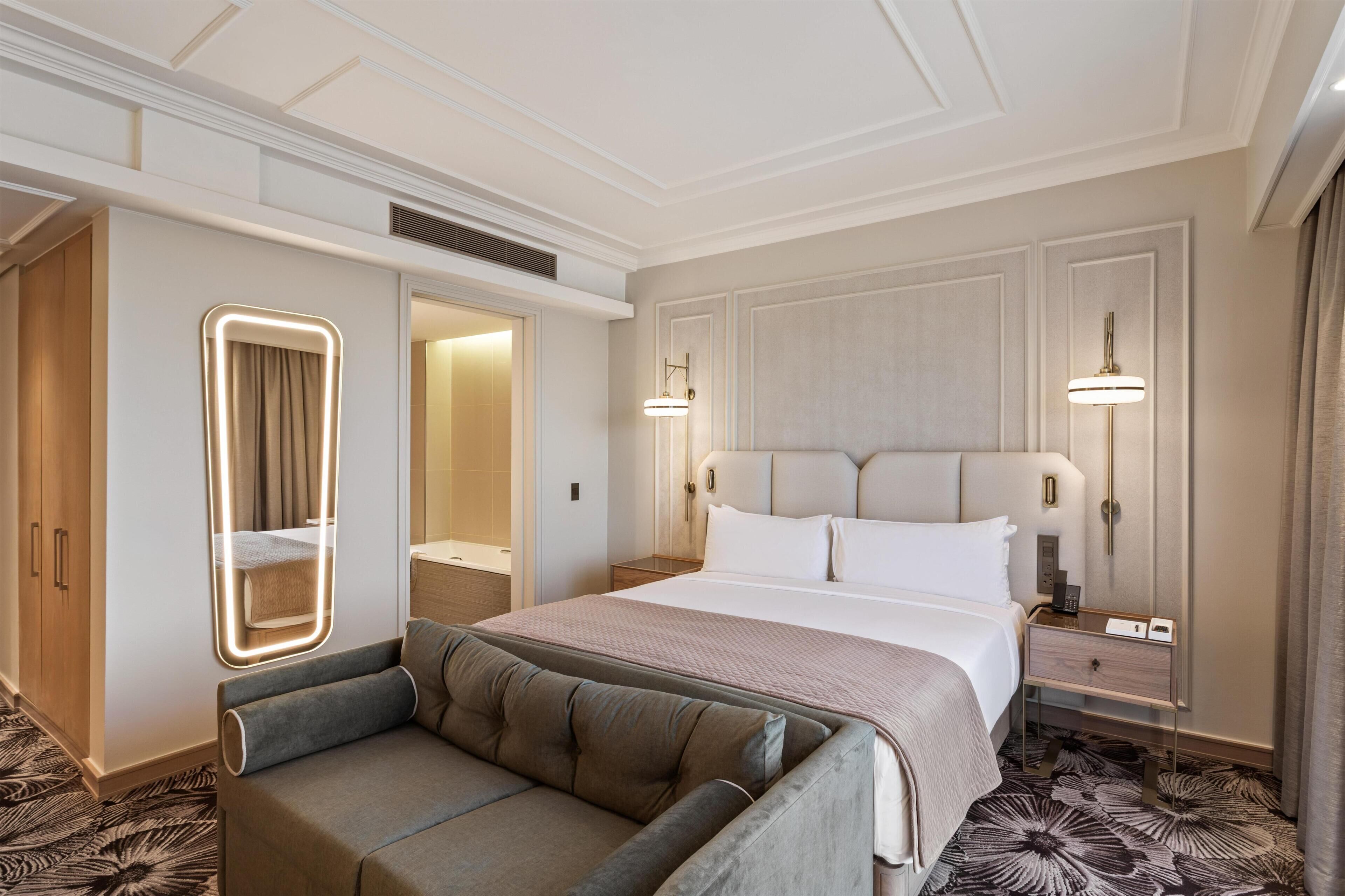 A hotel room with a bed, a sofa, a mirror, and a nightstand with a lamp.