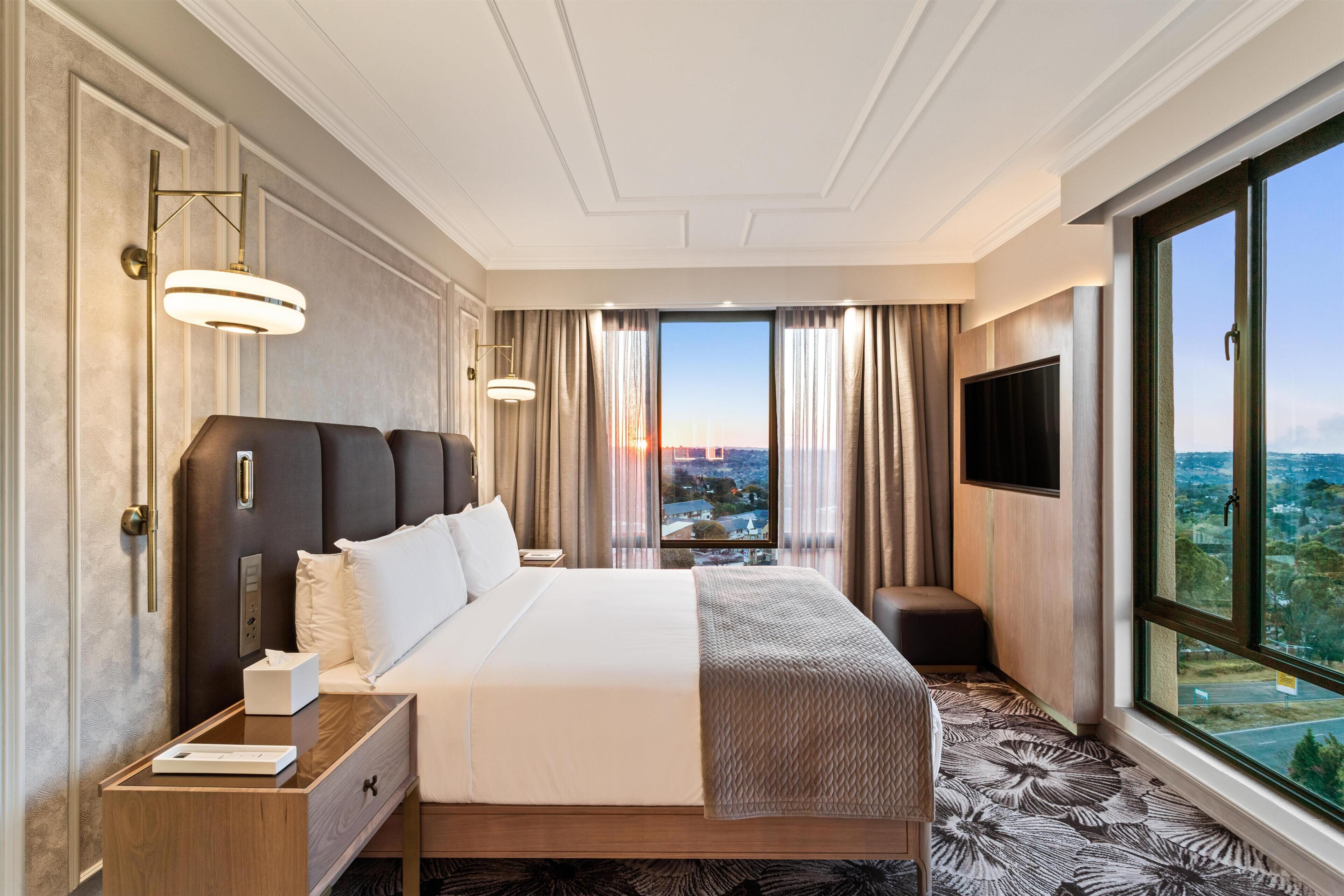 A hotel room with a large bed, a bedside table with a lamp, a TV, and a view of the city through the window.