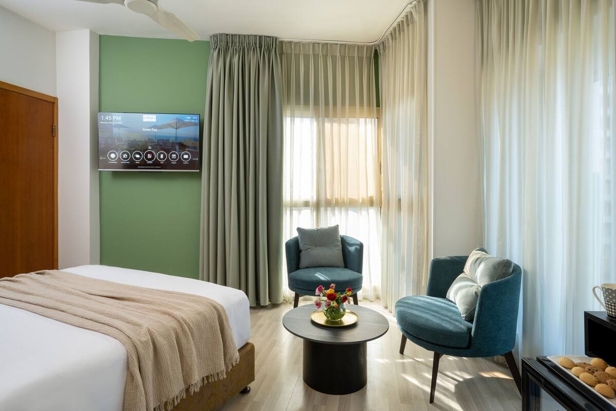 Habitación de hotel con cama, dos sillones, una mesita de centro con flores, televisor en la pared y vistas al exterior a través de cortinas translúcidas.