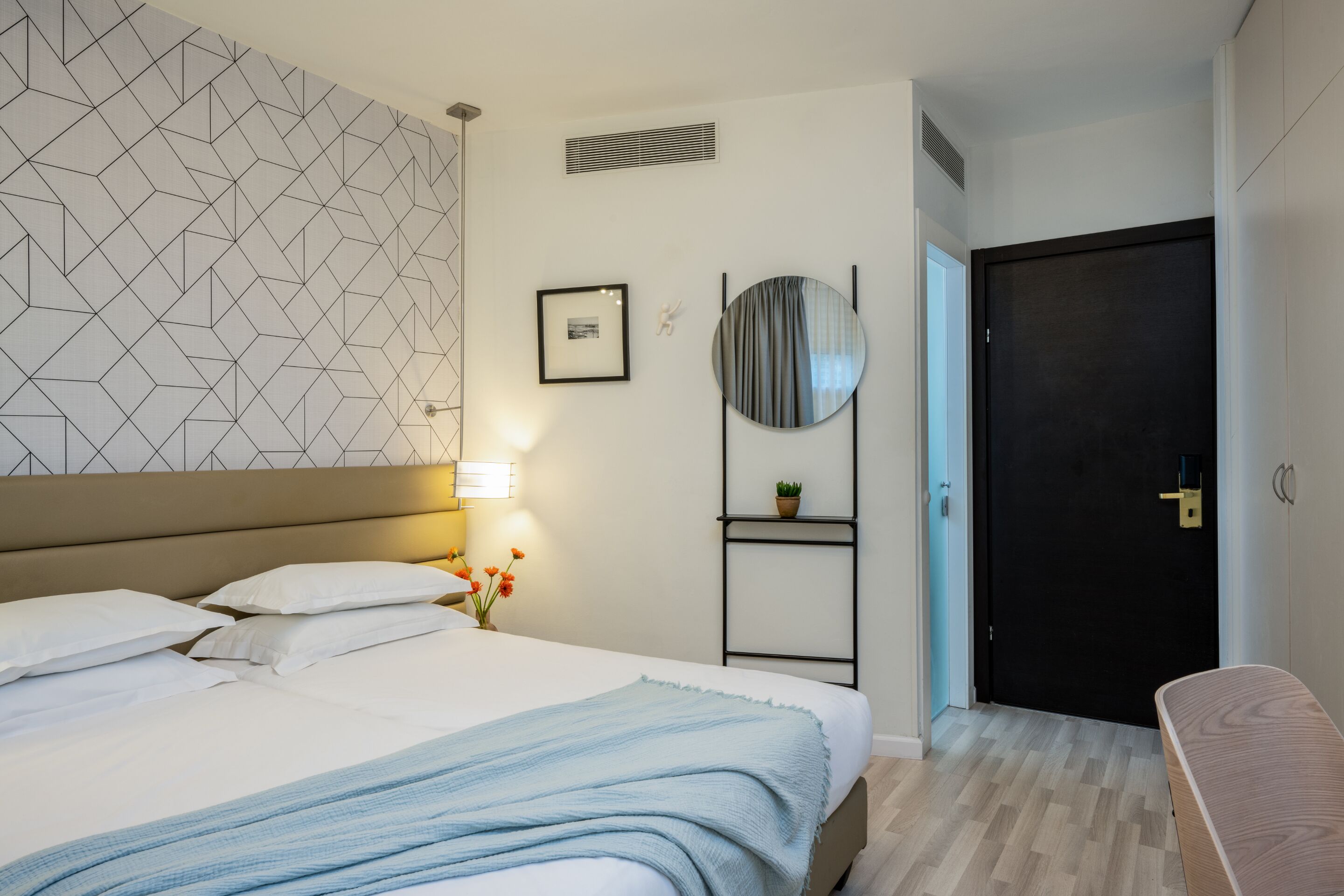 A hotel room with a bed, a bedside table with a lamp, a wall-mounted shelf with a plant, a door, and a wall with geometric patterns.