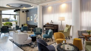 A modern hotel lobby with a piano, comfortable seating, and a large window offering a view of the outdoors. - Maxim Hotel Tel Aviv (Tel Aviv)
