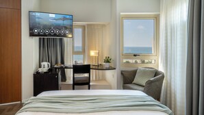 A hotel room with a bed, a desk with a TV, a chair, a small table with a lamp, and a view of the sea through a window. - Maxim Hotel Tel Aviv (Tel Aviv)