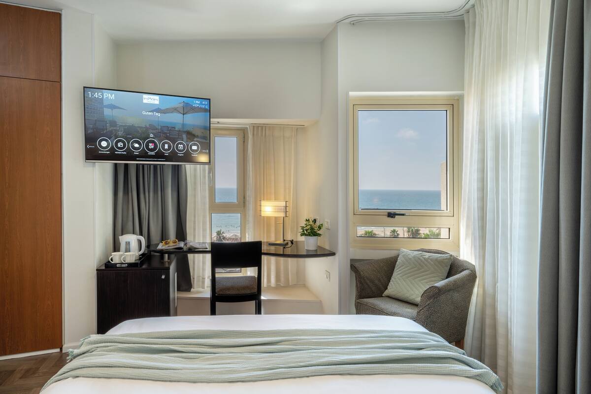 Habitación de hotel con cama, escritorio con televisor, silla, mesita de noche con lámpara y vista al mar por la ventana.