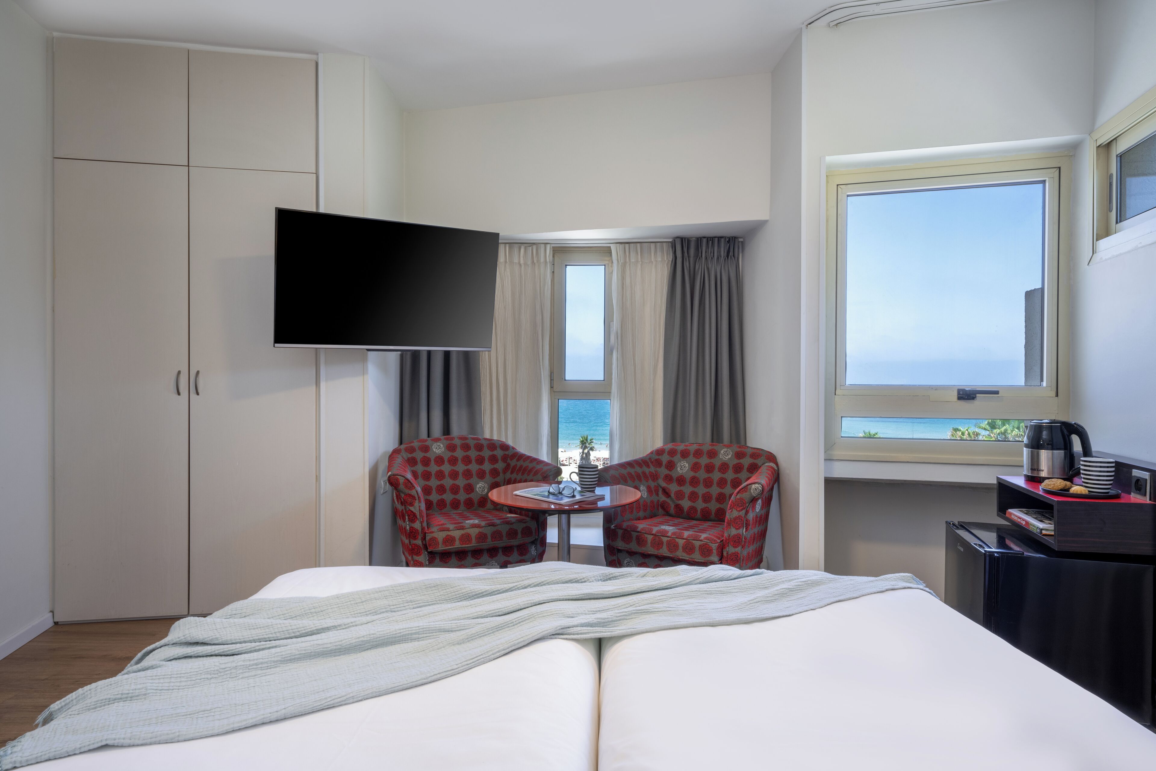 Ein Hotelzimmer mit einem Bett, einem Fernseher, einem Tisch mit zwei Stühlen, einem Fenster mit Blick auf den Strand und einer Minibar.