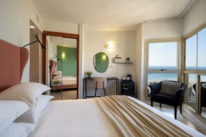 A hotel room with a large bed, a desk with a chair, a small table with a plant, a round mirror, and a view of the beach through large windows. - Maxim Hotel Tel Aviv (Tel Aviv)