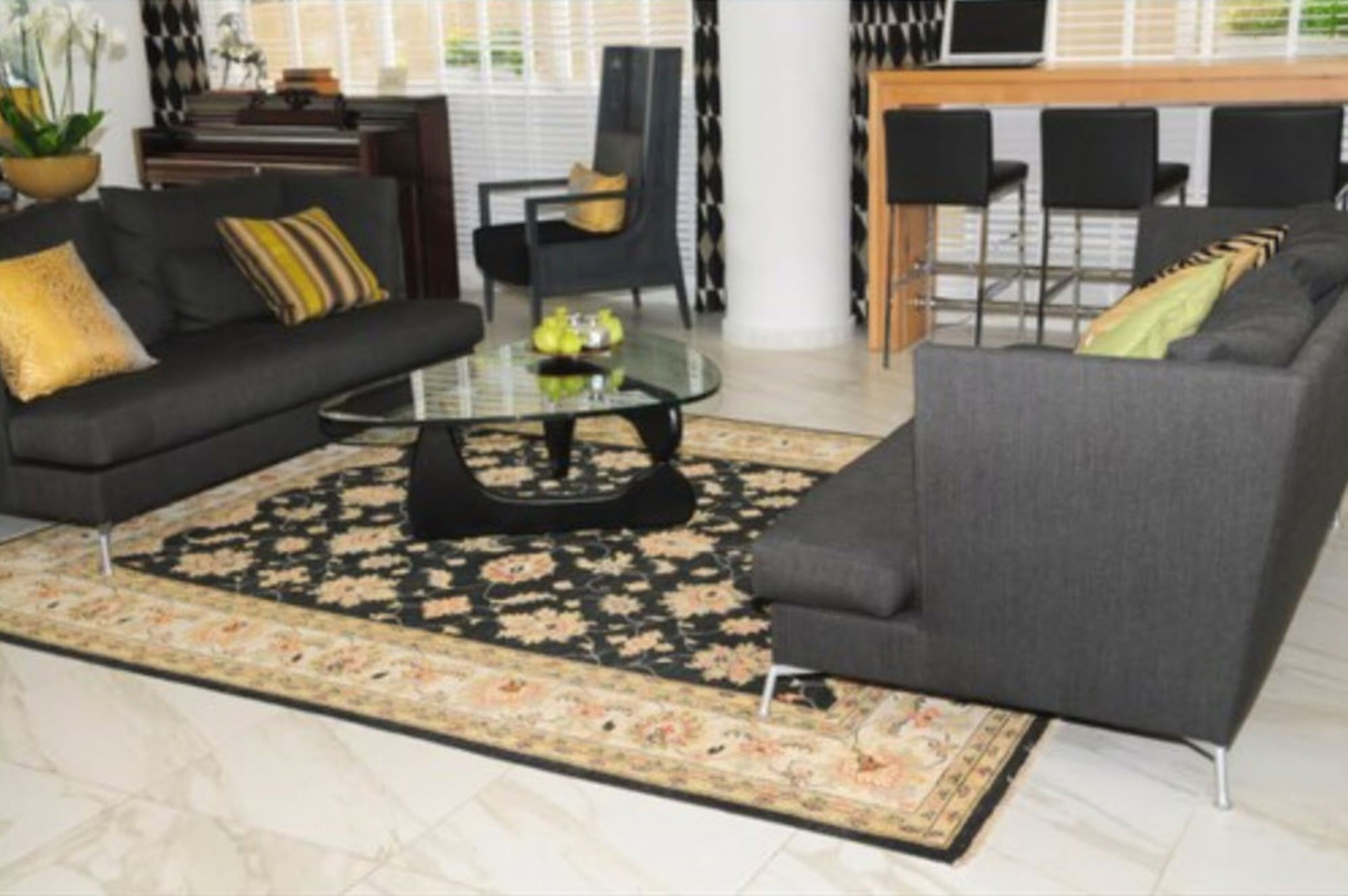Una sala moderna con un sofá gris oscuro, una mesa de centro de vidrio y un tapete estampado. Hay un comedor con barra y sillas negras.
