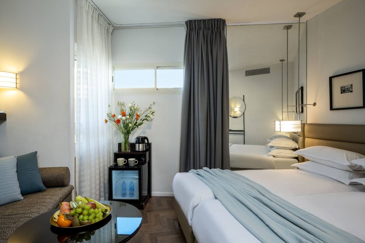 Habitación de hotel con cama, sofá, una mesita con frutero, mesita de noche con lámpara y ventana con cortinas.