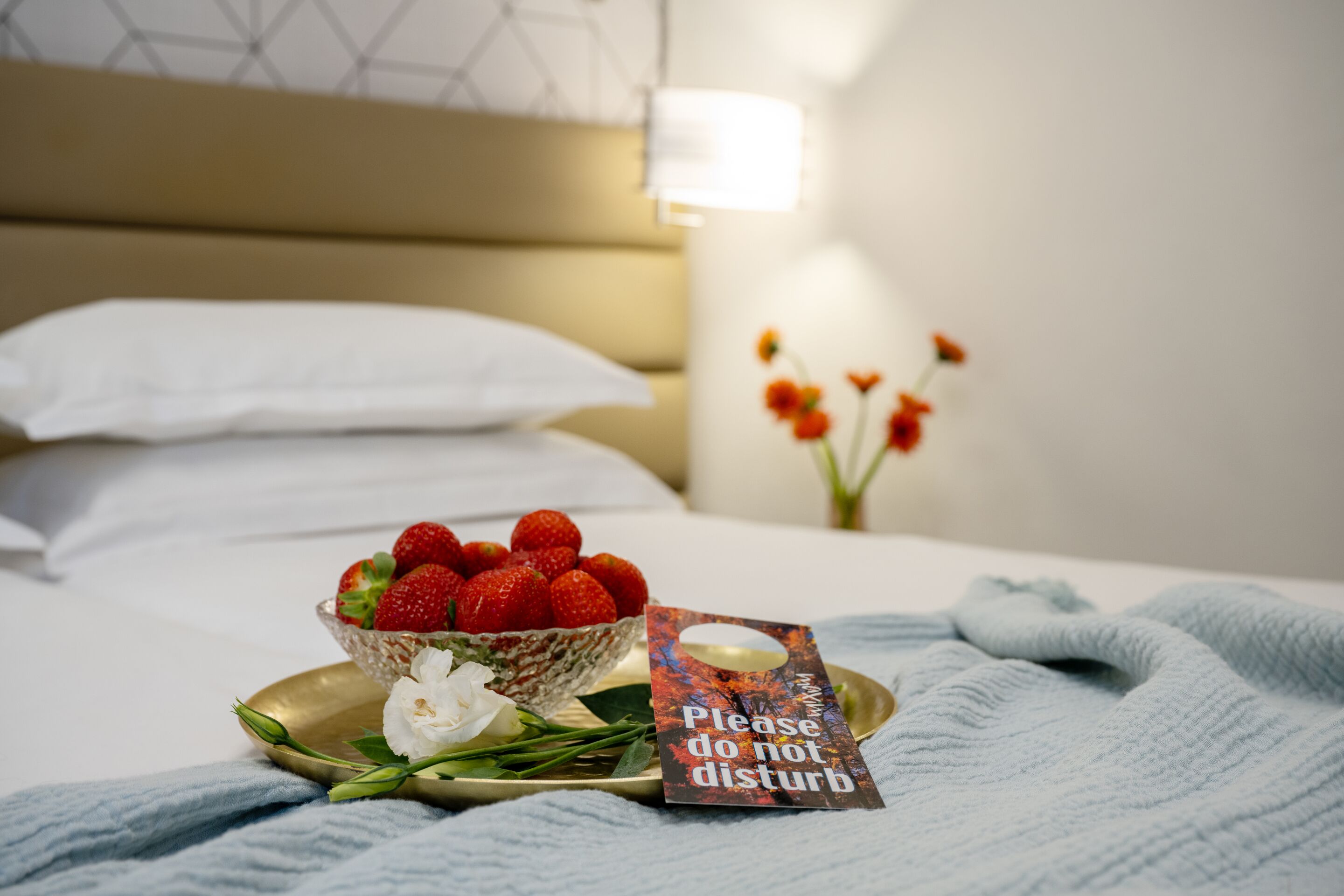 A neatly arranged breakfast tray with strawberries, a white flower, and a 'Please do not disturb' sign on a bed in a hotel room.