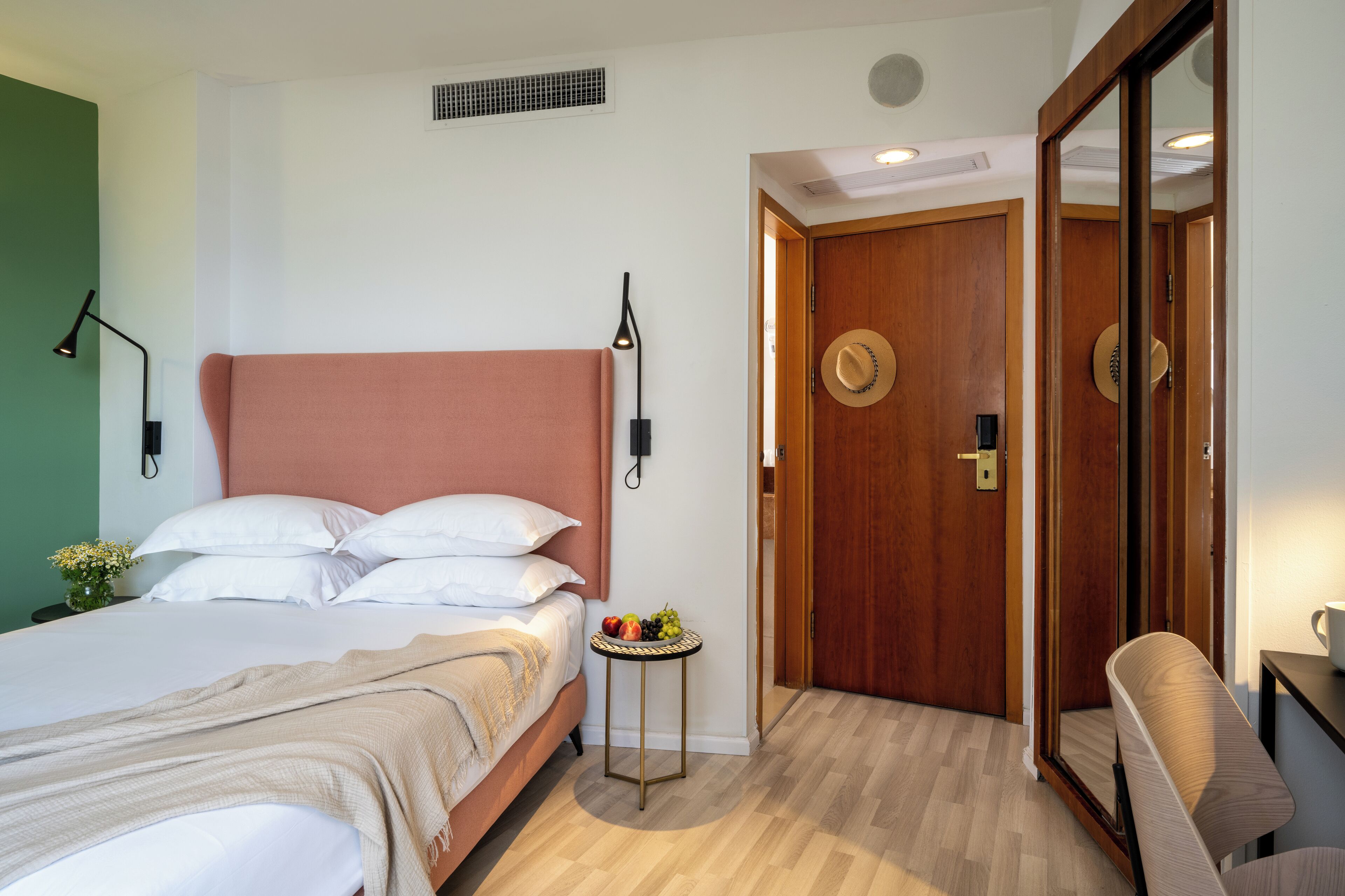 Een hotelkamer met een bed, nachtlampjes, een kleintje tafeltje met fruit, een spiegel en een deur.