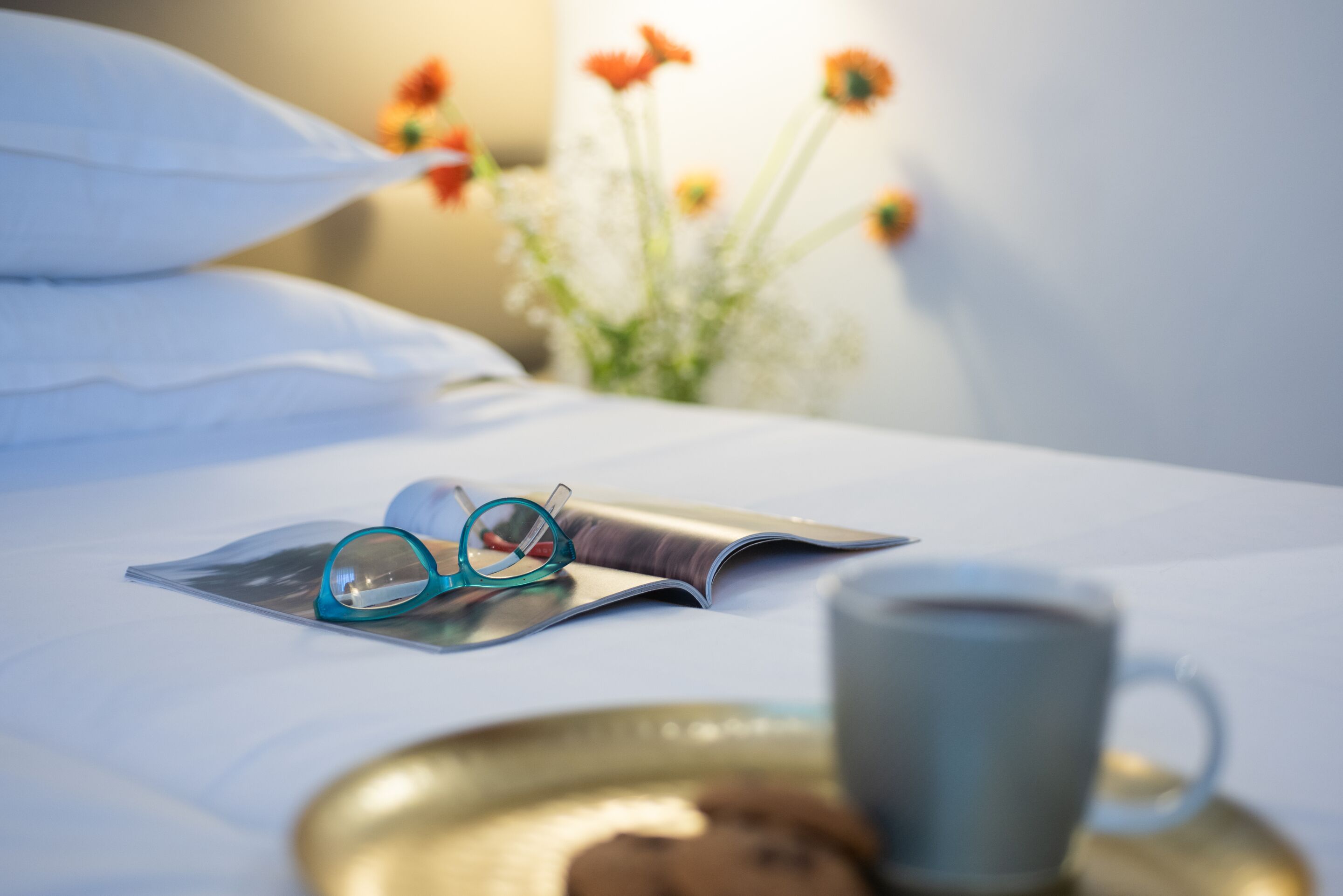 A neatly made bed with a pair of glasses, a magazine, and a cup on a tray.