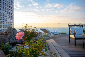 A wooden deck with lounge chairs and a table, overlooking a body of water and a building. - Maxim Hotel Tel Aviv (Tel Aviv)