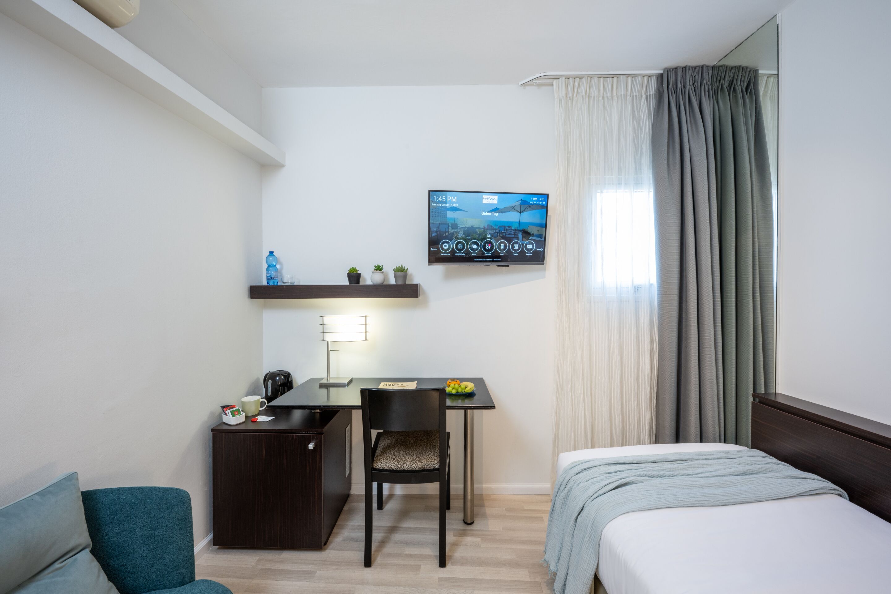 Une chambre d’hôtel comprenant un lit, un bureau avec une chaise, un téléviseur fixé au mur et une fenêtre avec des rideaux.