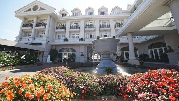 A grand hotel with a central fountain, surrounded by vibrant flower beds, and multiple balconies with railings. - Sammy Dalat Hotel (Xuan Huong - Da Lat)