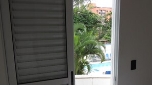 Een kamer met een bed, een raam met zonwering en uitzicht op een zwembad en palmbomen.