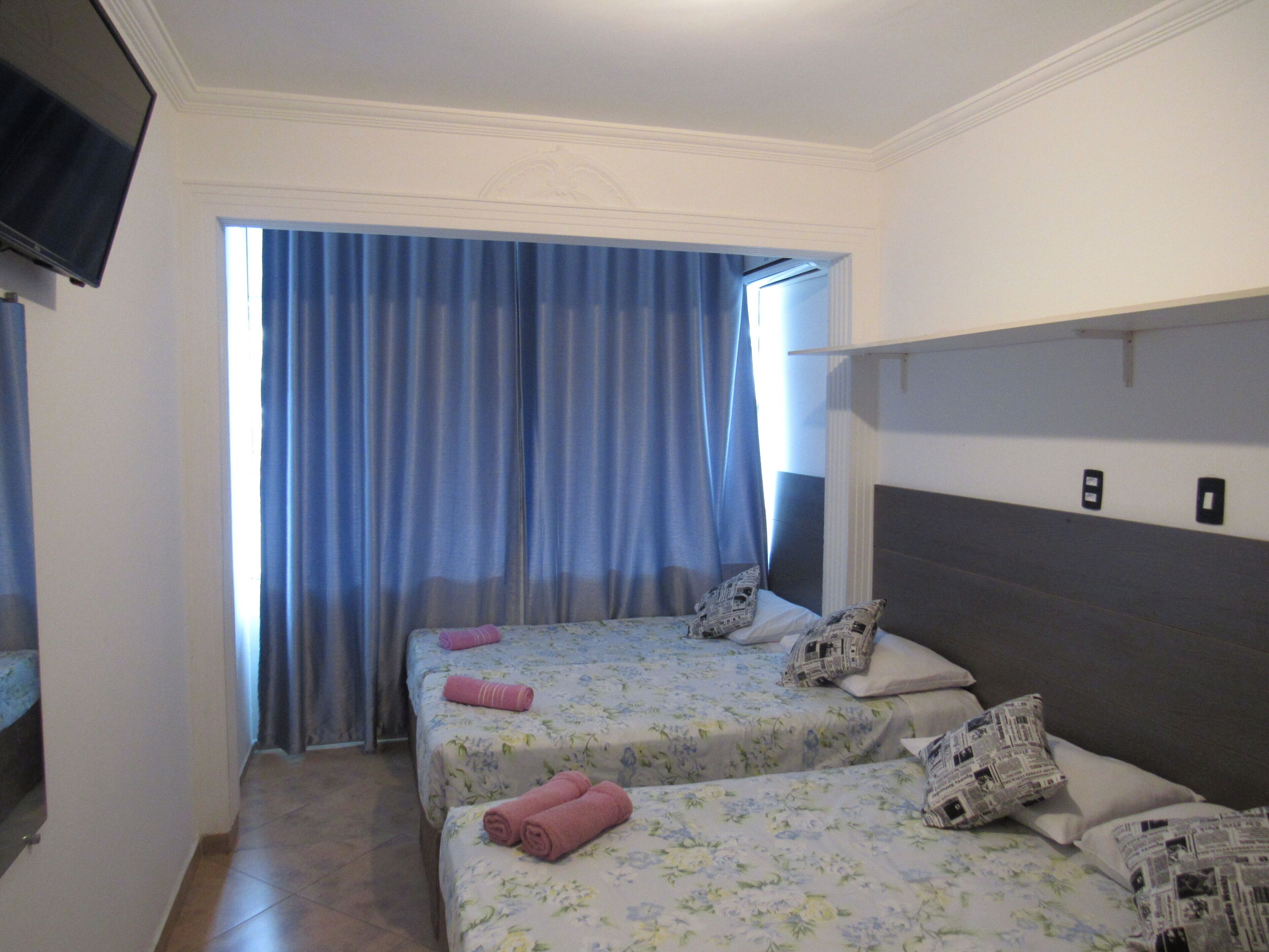 A hotel room with two beds, each with floral bedding and pillows, a flat-screen TV mounted on the wall, and a large window with blue curtains.
