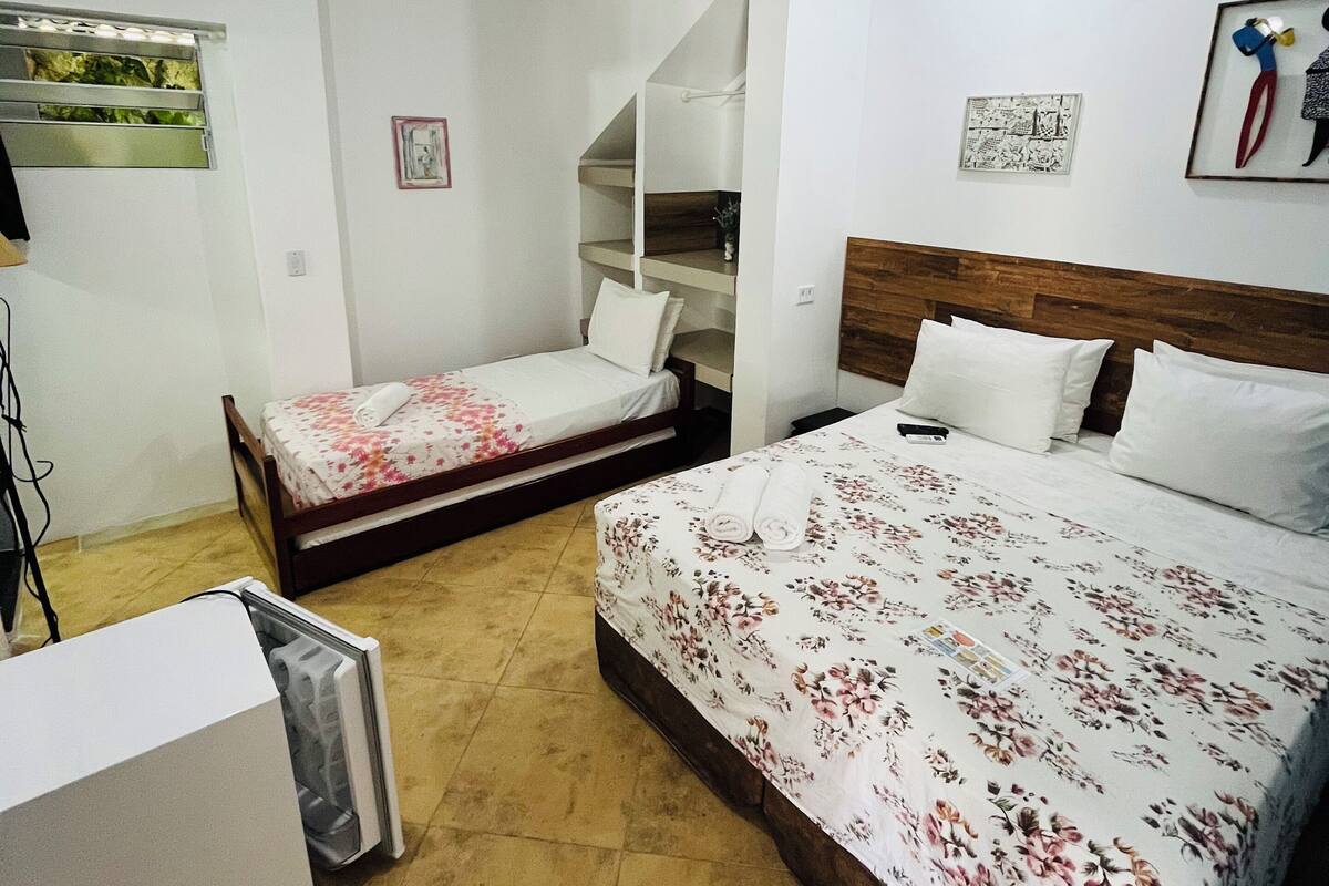Une chambre d’hôtel avec deux lits, une tête de lit en bois, un climatiseur blanc, une petite table et une étagère fixée au mur.