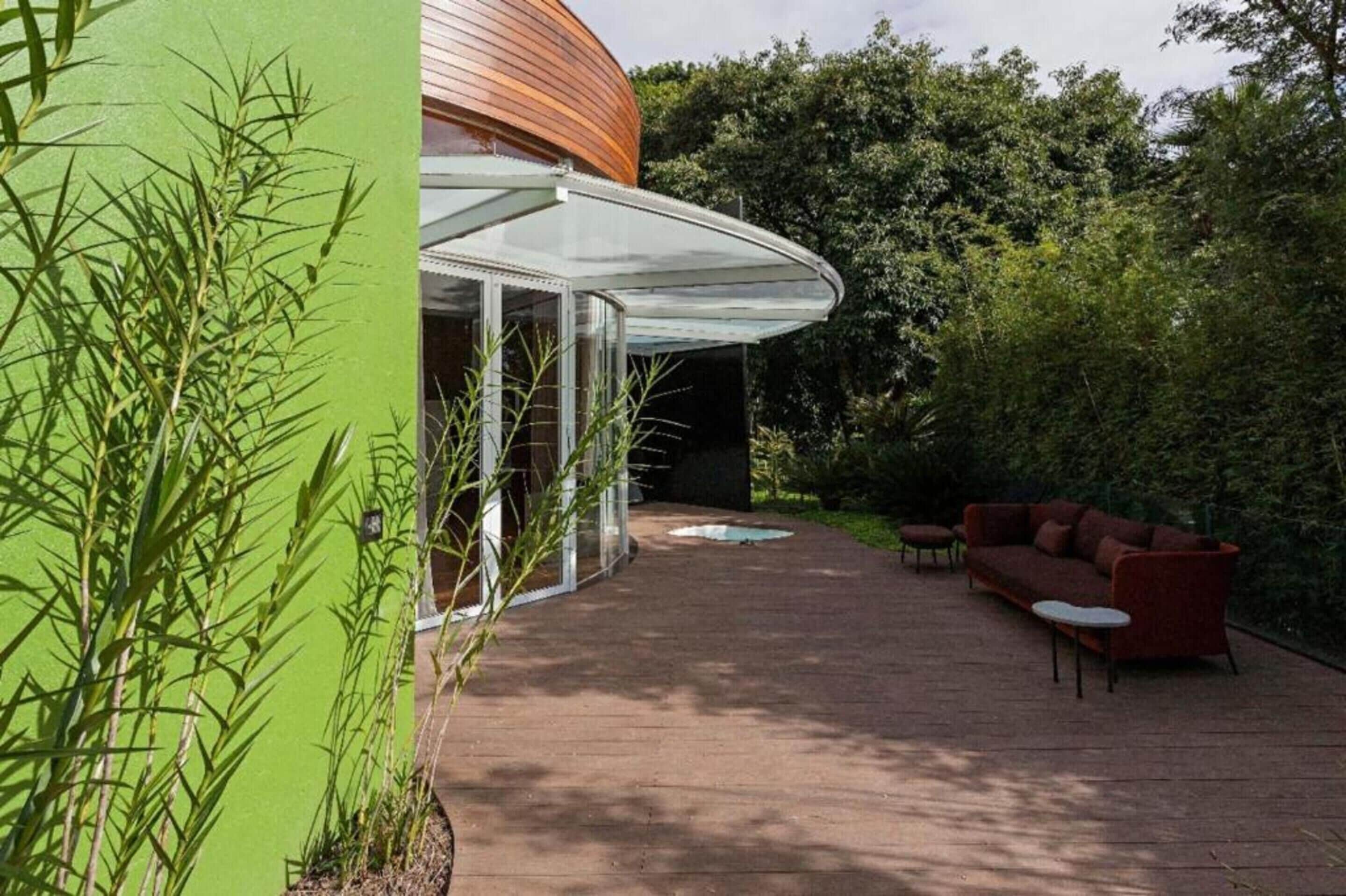 A modern outdoor patio with a curved wooden structure, a glass enclosure, and a seating area with a red sofa and table.
