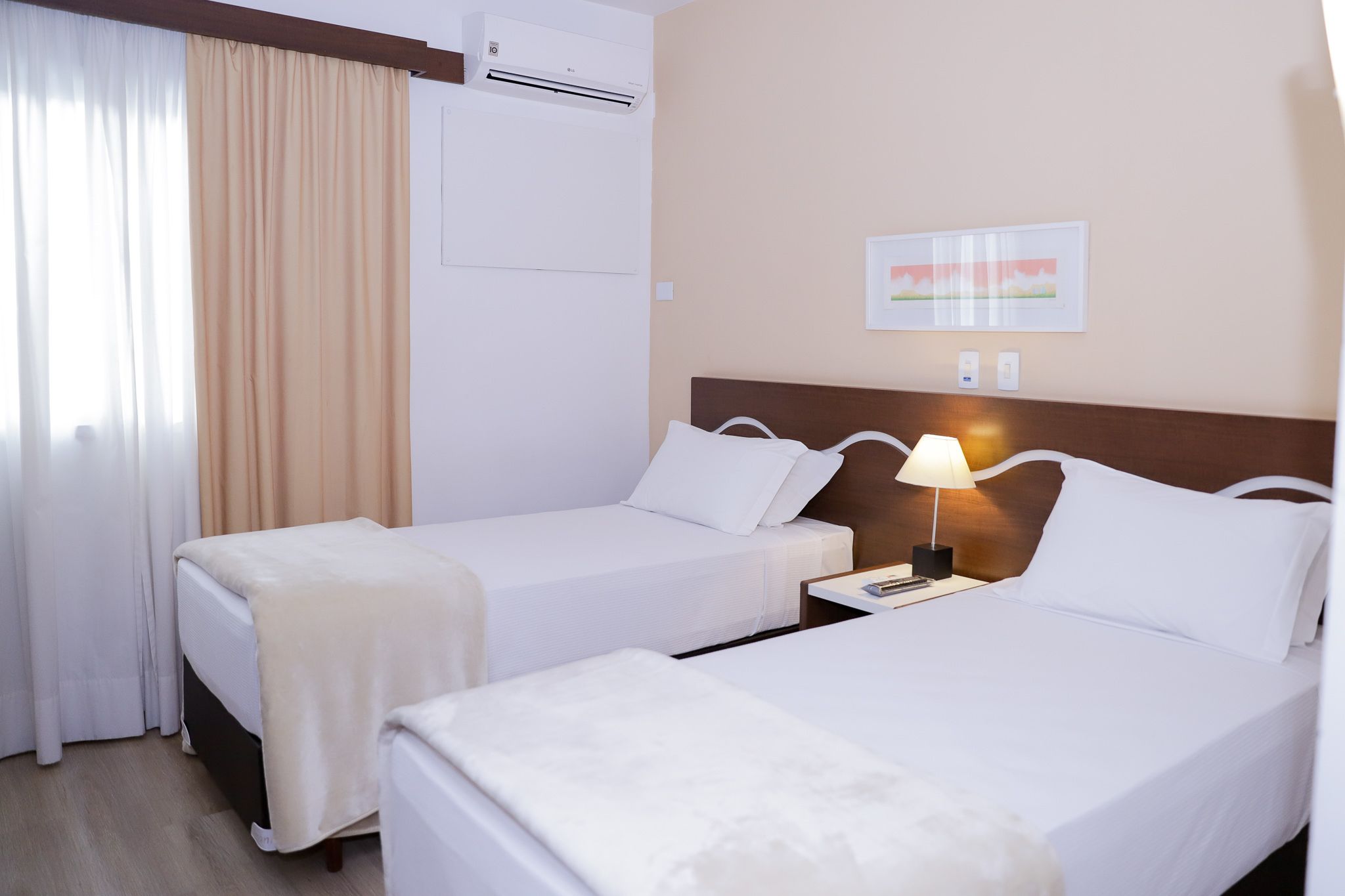 Una habitación de hotel con dos camas, una mesita de noche con lámpara, un equipo de aire acondicionado y un cuadro enmarcado en la pared.