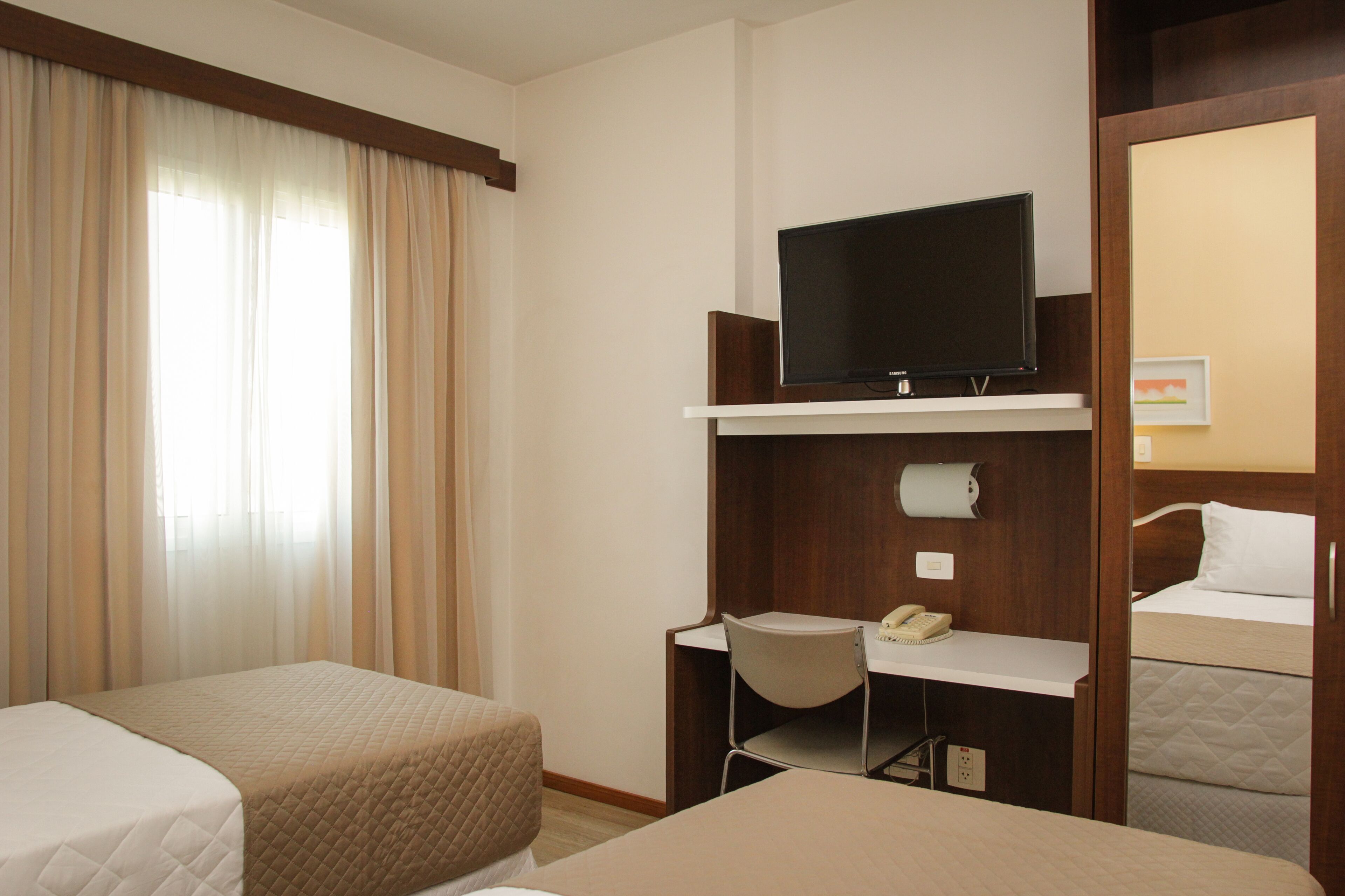 A hotel room with two beds, a desk with a telephone, a television on a shelf, and a window with curtains.