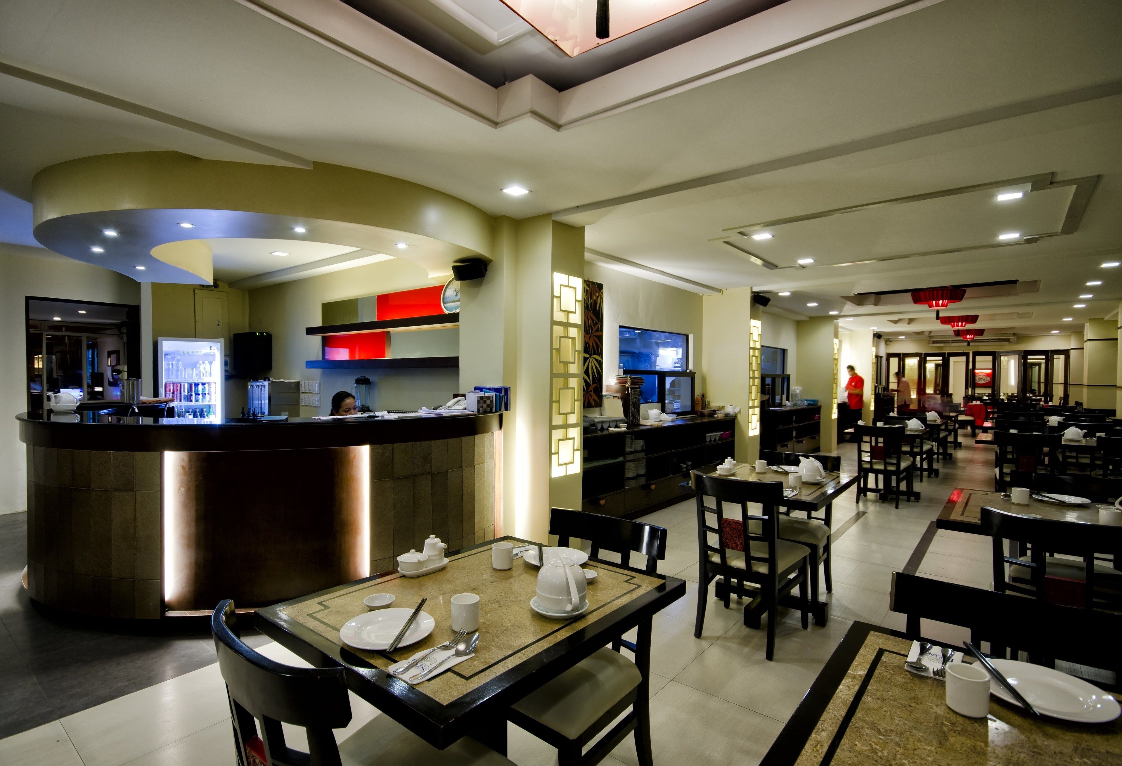 A spacious restaurant with multiple dining tables and chairs, a counter with a beverage station, and a television screen.