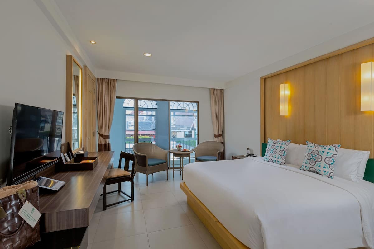 Habitación de hotel con una cama grande, un escritorio con televisor, dos sillas, una mesita y vistas a la ciudad a través de amplias ventanas.