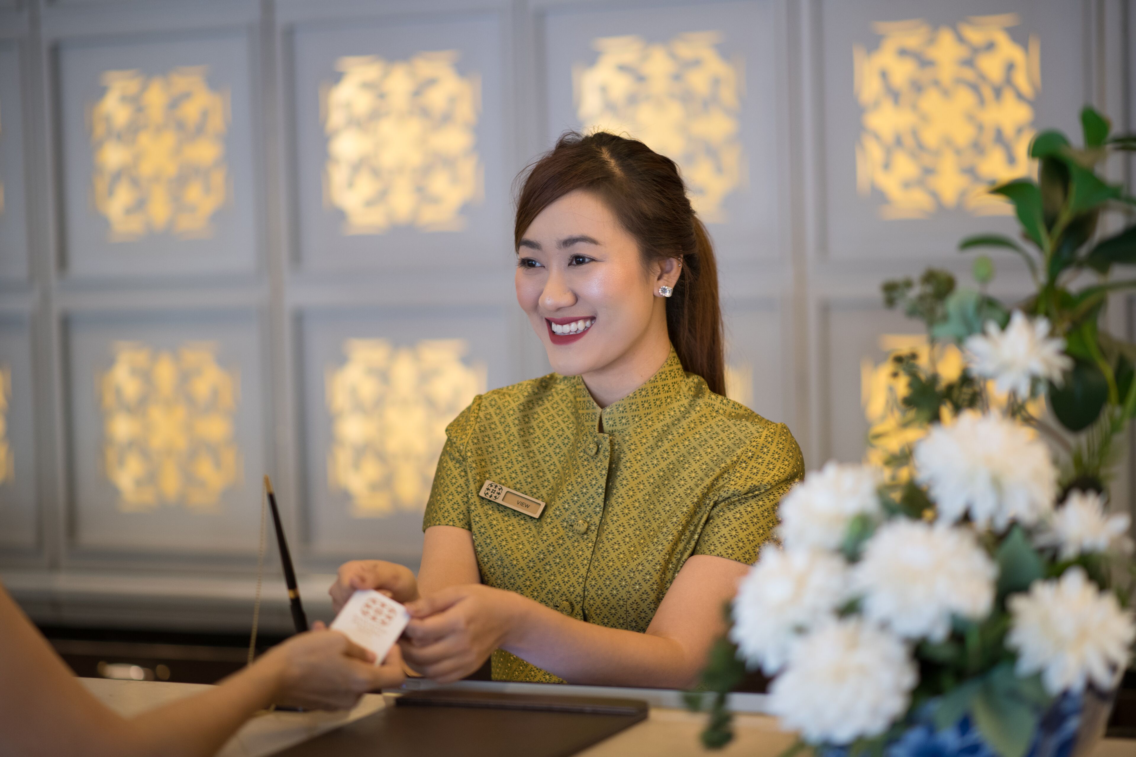 Un'area reception con una receptionist, un cartellino identificativo, un tablet e un'esposizione floreale.