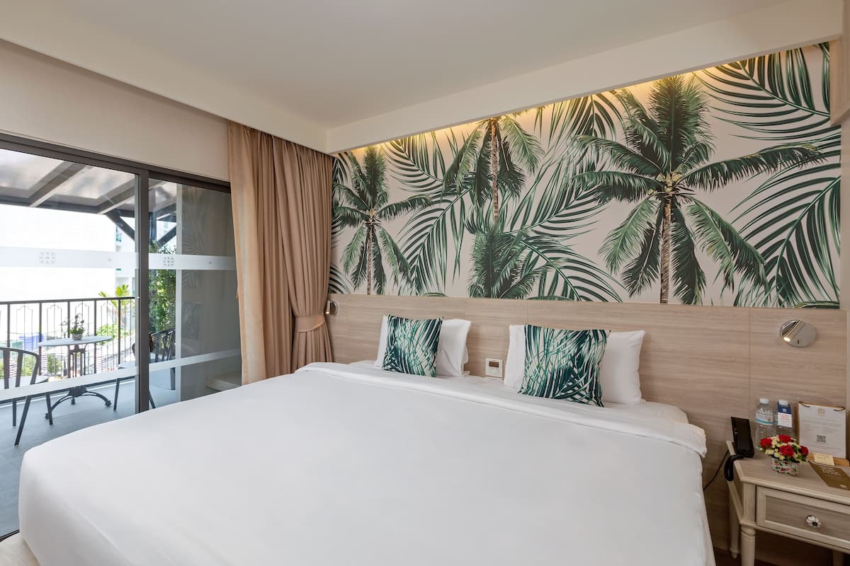 Una habitación de hotel con una cama grande, una mesita de noche con un teléfono y un jarrón de flores, una pared con un mural de palmeras y un balcón con una mesa y sillas.