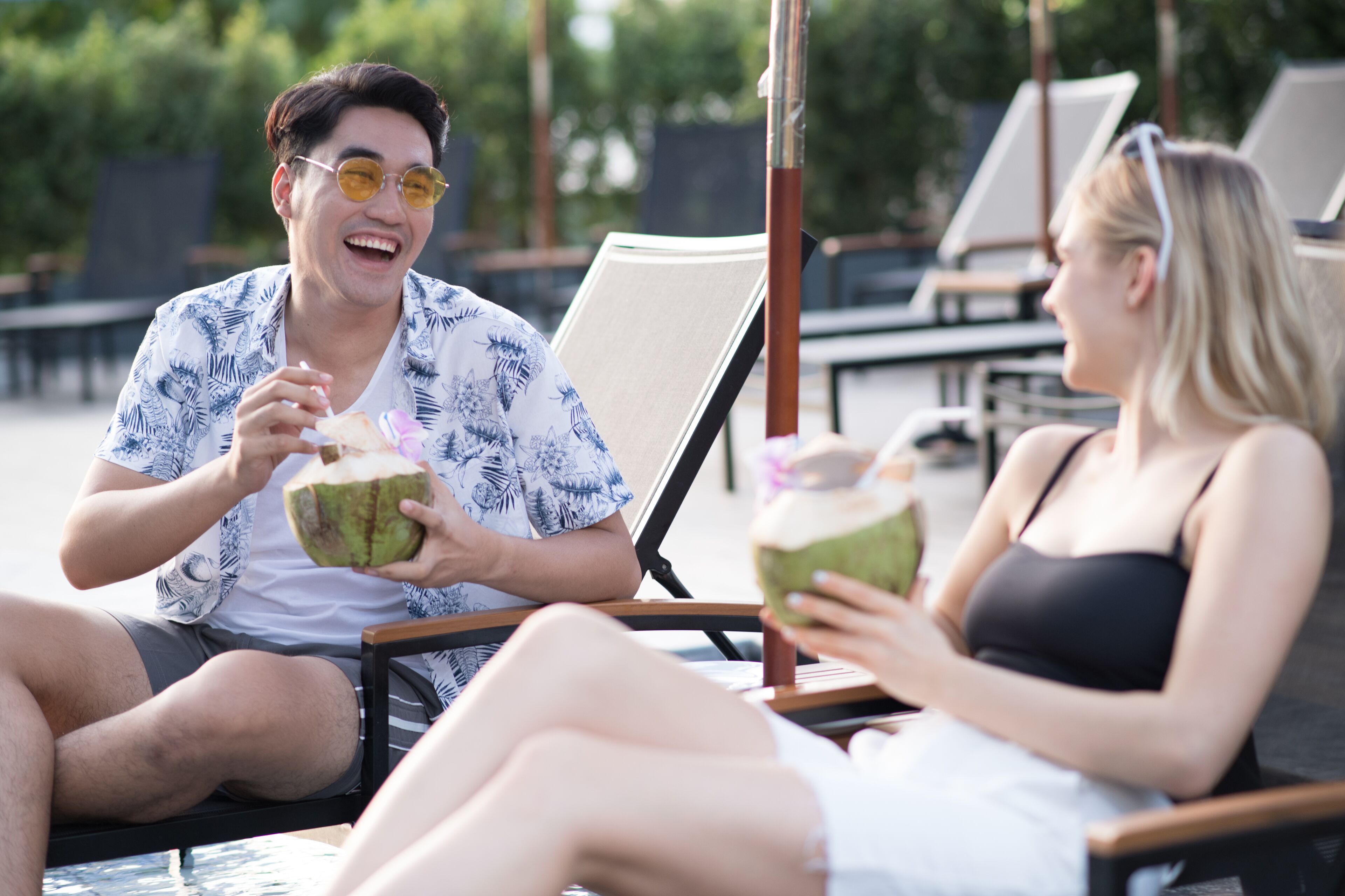 Two people sitting on lounge chairs by a pool, holding coconuts.