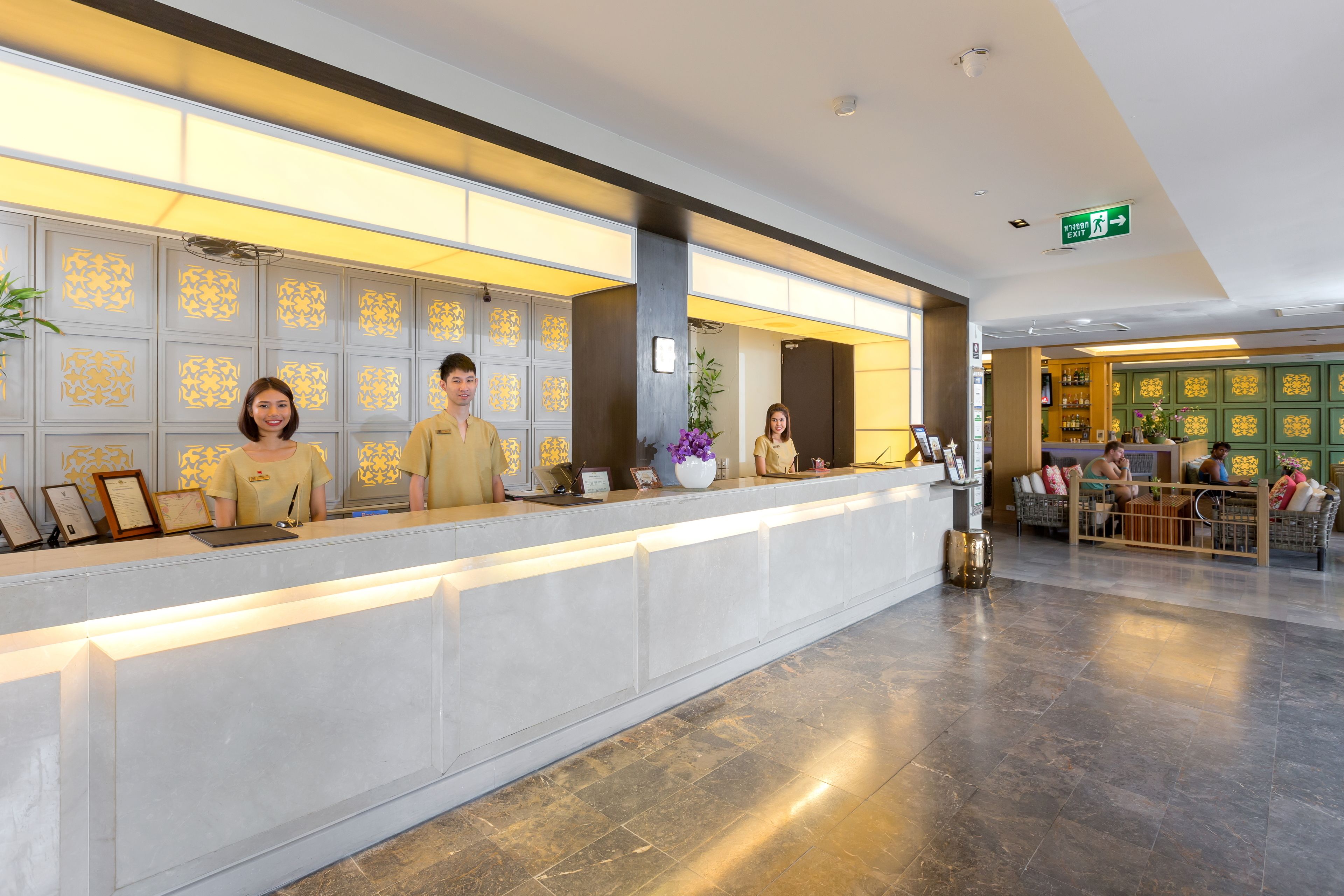 A hotel reception area with a modern design, featuring a reception desk, a seating area with wicker furniture, and a decorative wall with patterned panels.
