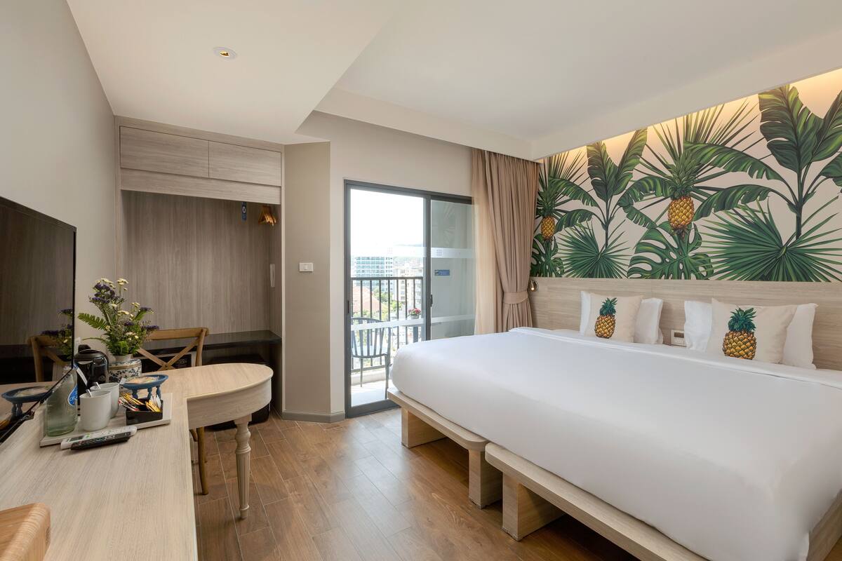 Habitación de hotel con una cama grande, una mesa de comedor, un televisor y un balcón con un mural tropical.