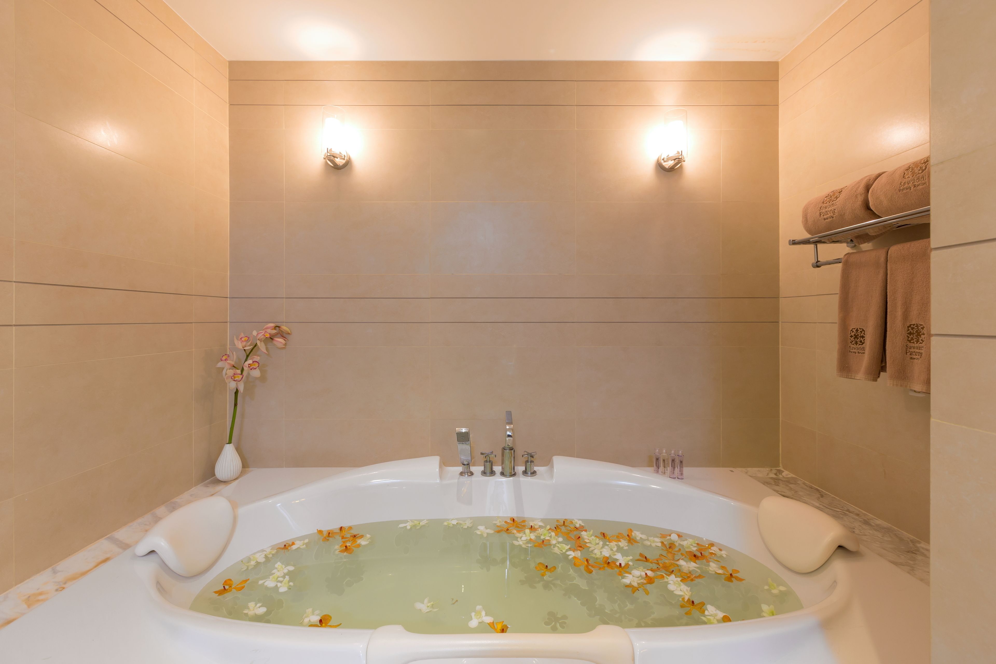 A modern bathroom with a large bathtub, beige tiles, and a vase of flowers.