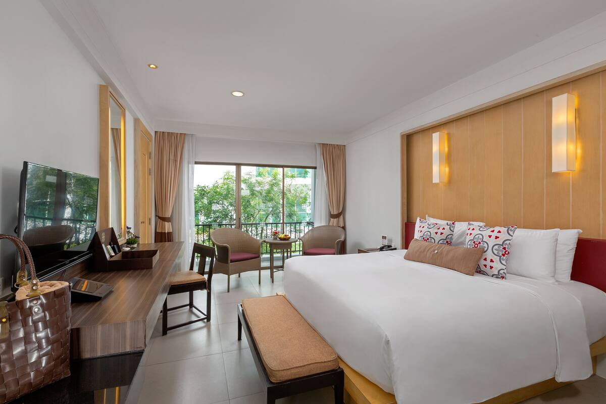 Habitación de hotel con una cama grande, un escritorio con televisor, una pequeña mesa con sillas y un balcón con cortinas.