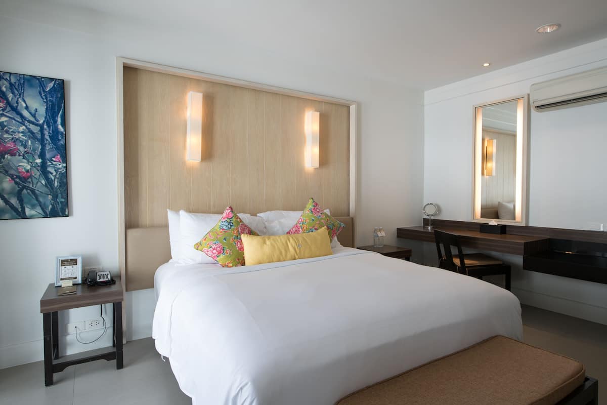 Una habitación de hotel moderna con una cama grande, un cabecero de madera, una mesita de noche con lámpara, un espejo y un pequeño escritorio con una silla.