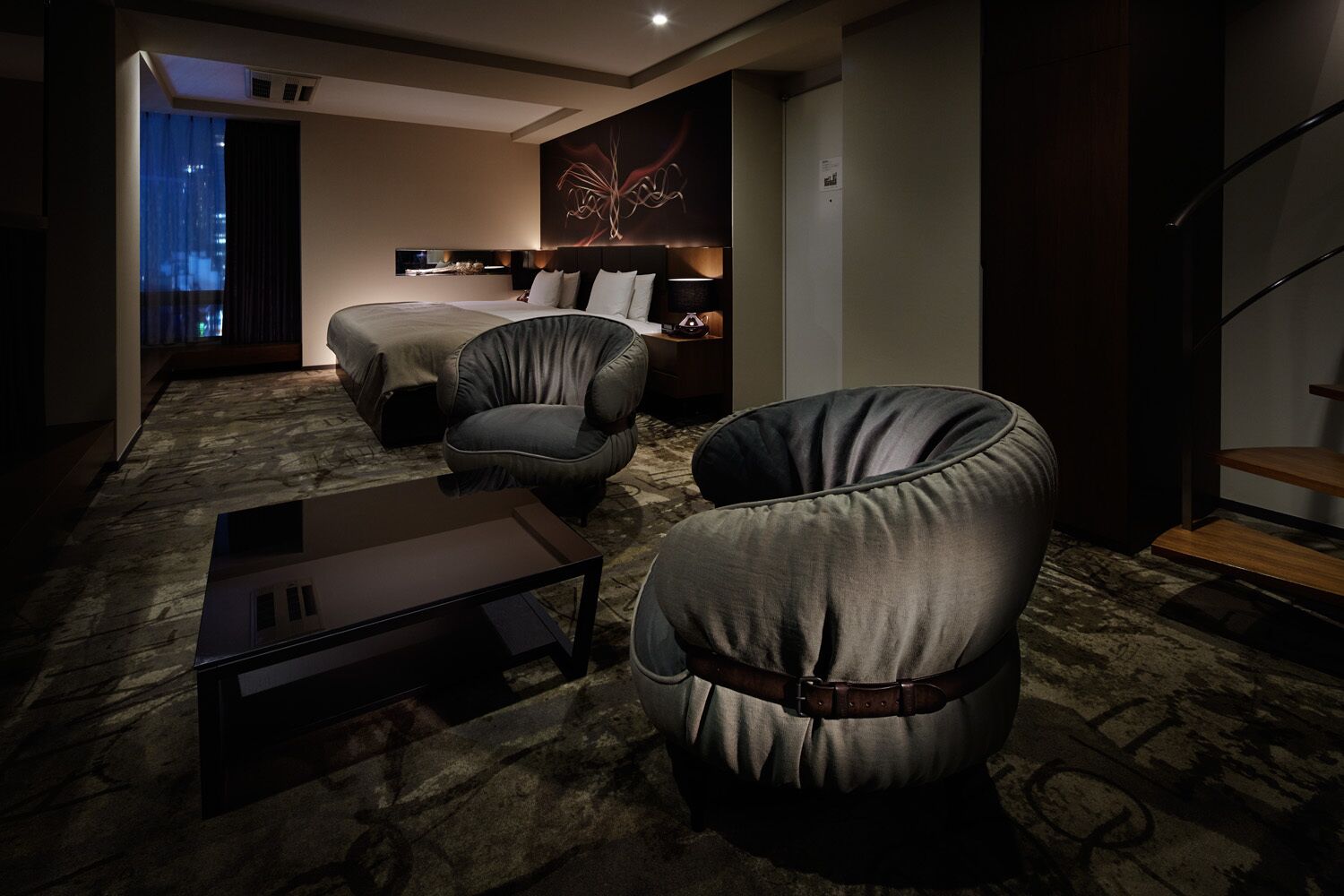 A hotel room with a bed, two armchairs, a small table, a wall art piece, and a staircase.