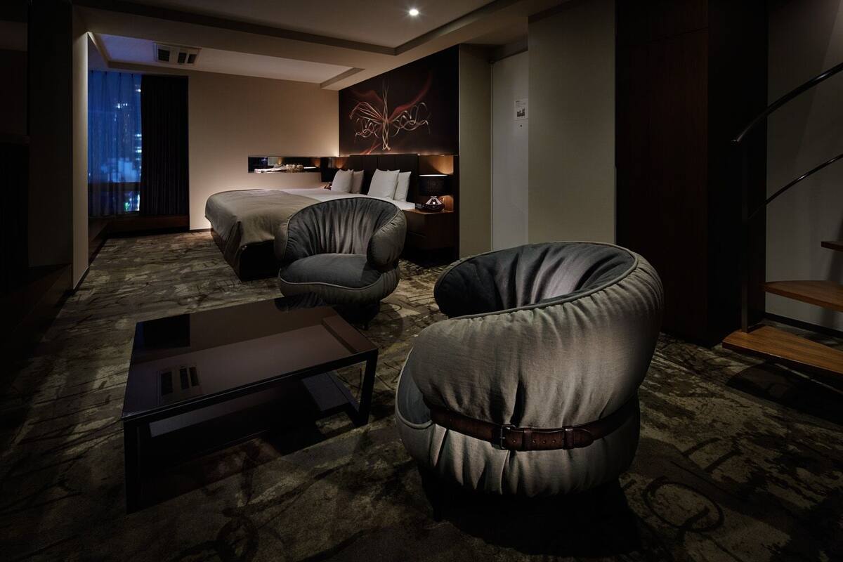 A hotel room with a bed, two armchairs, a small table, a wall art piece, and a staircase.