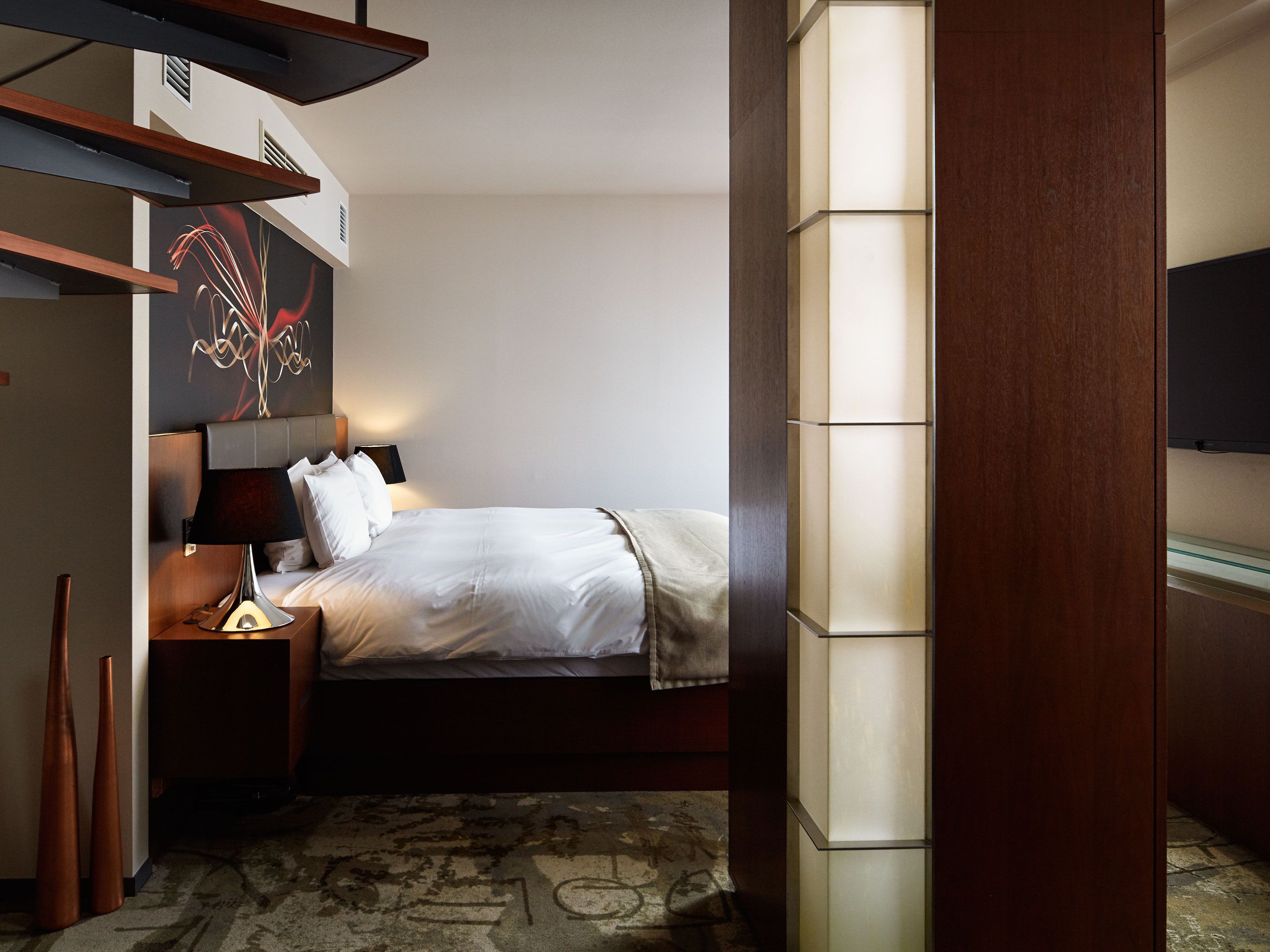 Una habitación de hotel con cama, mesitas de noche, lámpara, una obra de arte en la pared y un ventanal con cortinas.