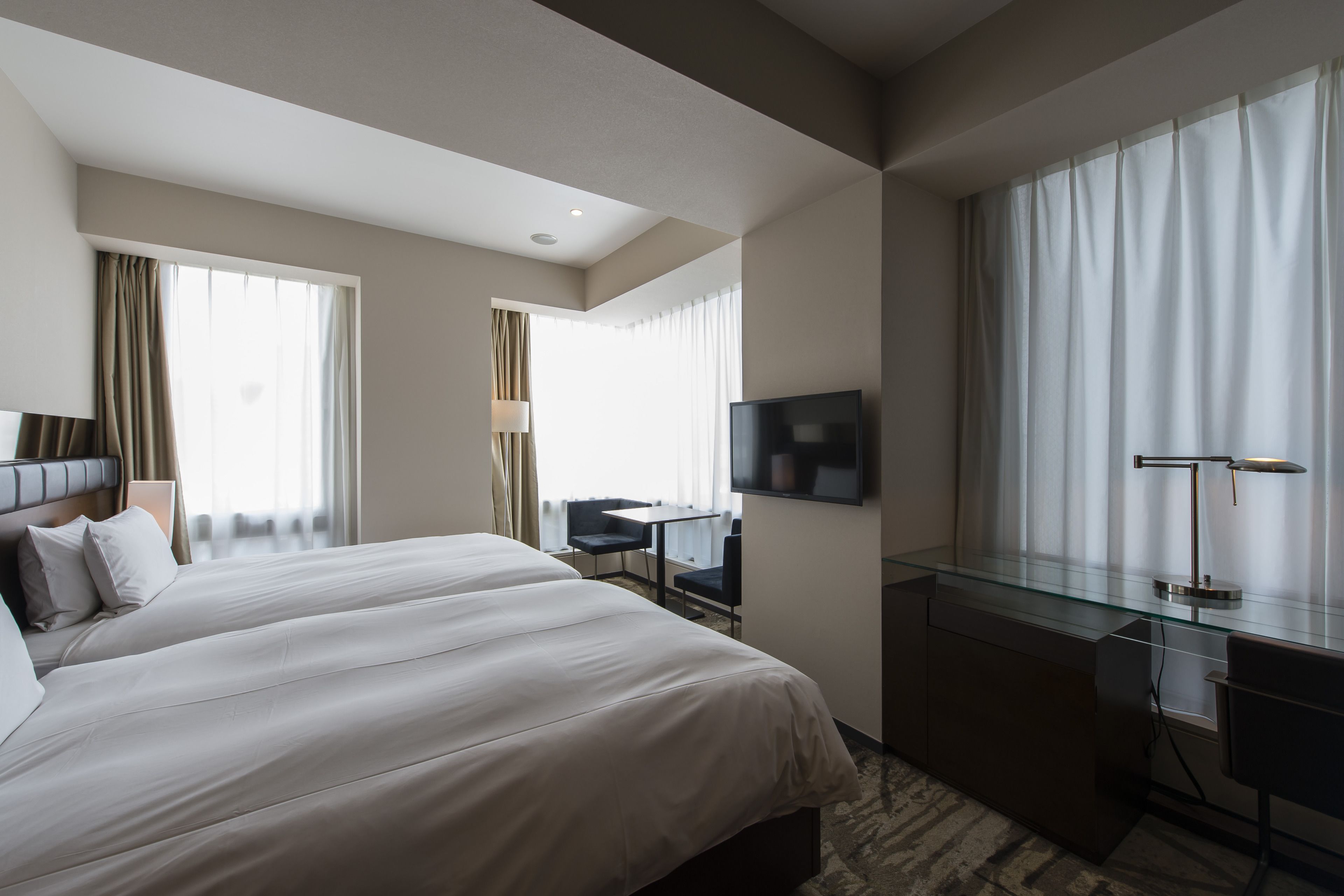 A hotel room with two beds, a desk, a chair, a TV, and large windows with curtains.