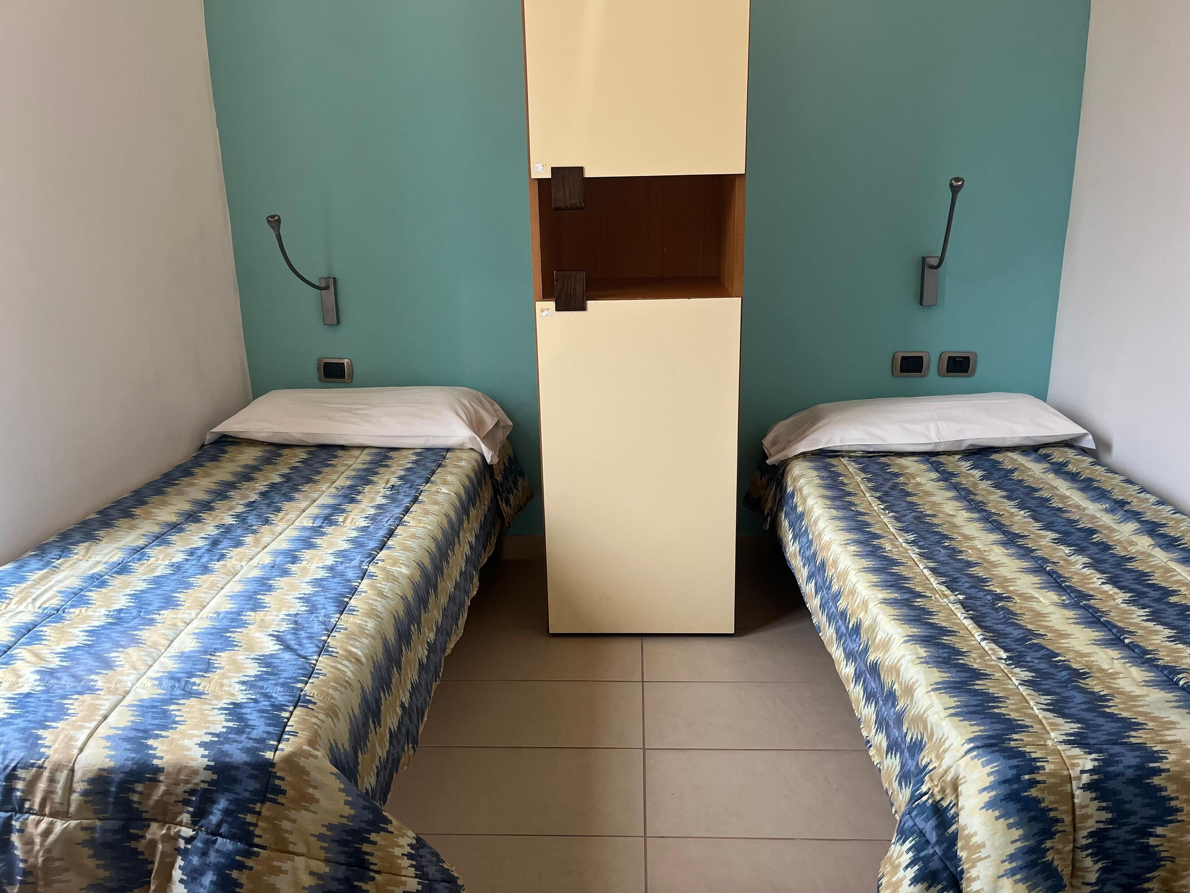 A room with two single beds, each with a patterned bedspread, a central partition, and a wall-mounted light fixture.