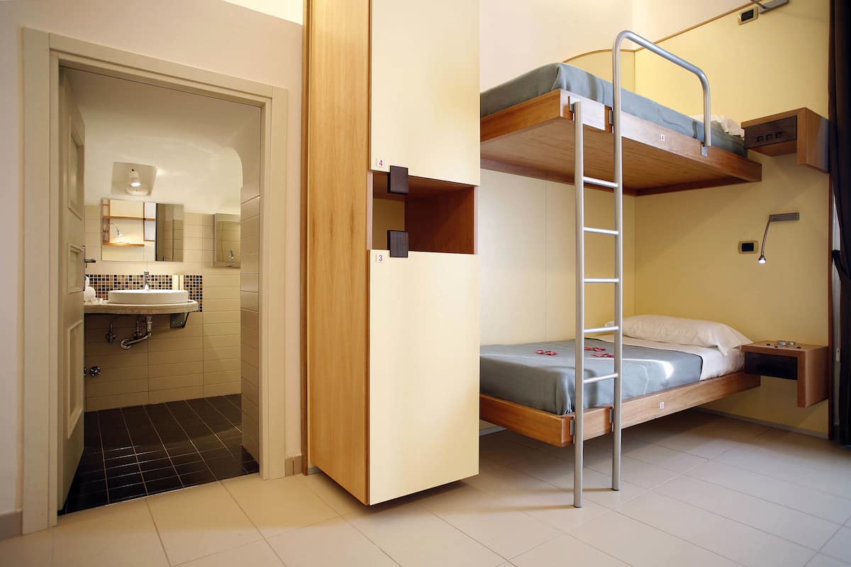 A compact room with a bunk bed, a small bedside table, a built-in shelf, and a bathroom with a sink and mirror.