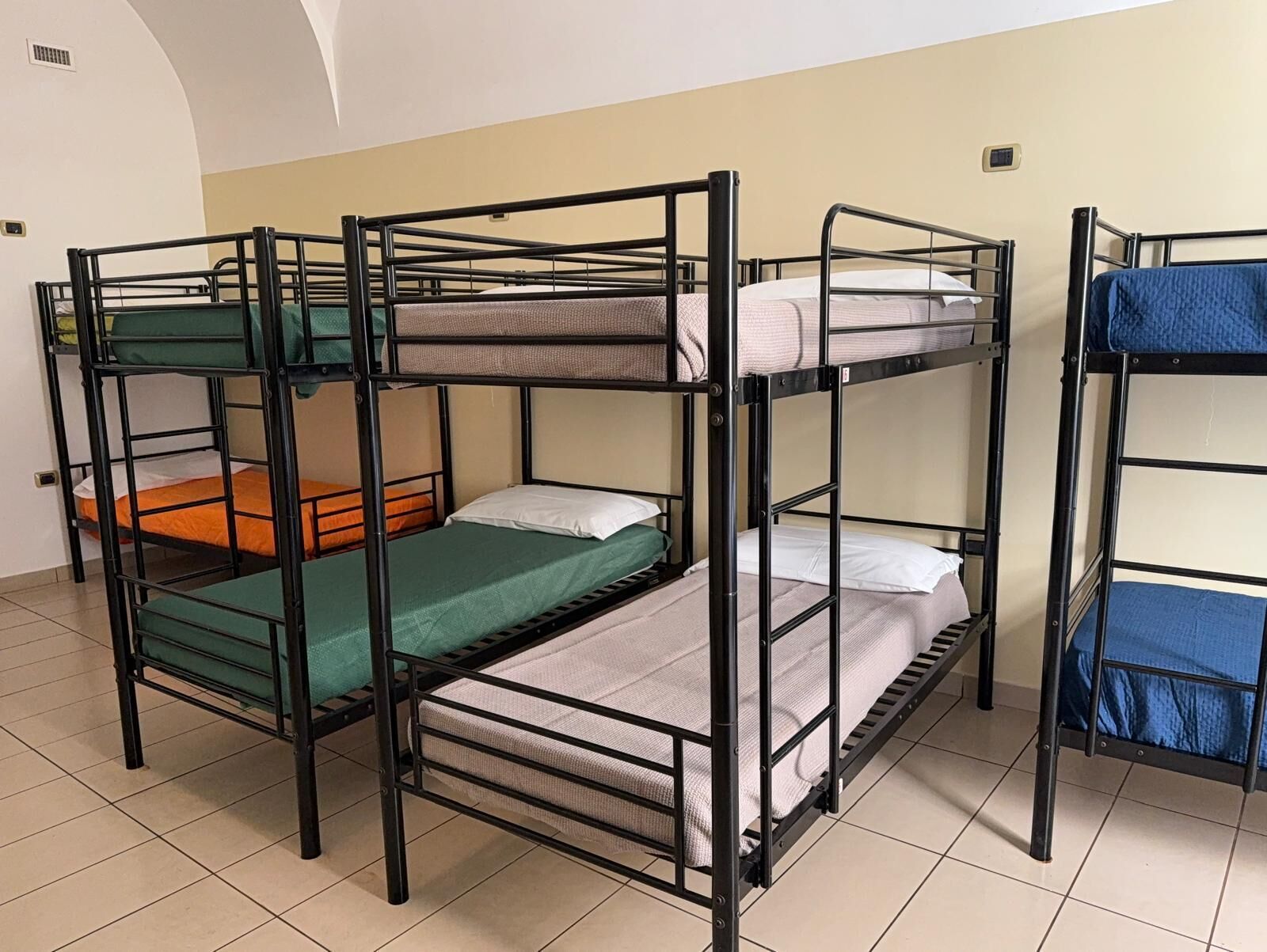 Habitación tipo dormitorio con literas, suelo de baldosas y una pared blanca con un respiradero.