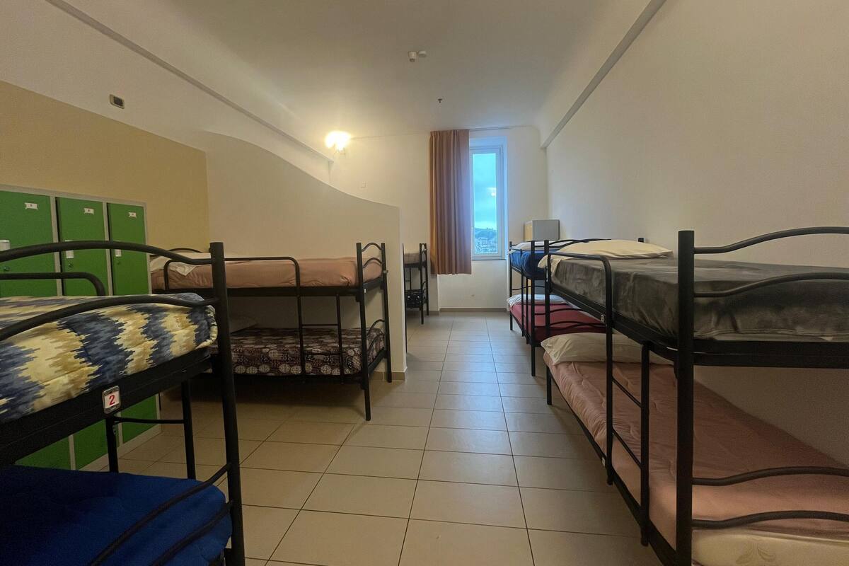 A dormitory room with bunk beds, a window with curtains, and green lockers in the background.