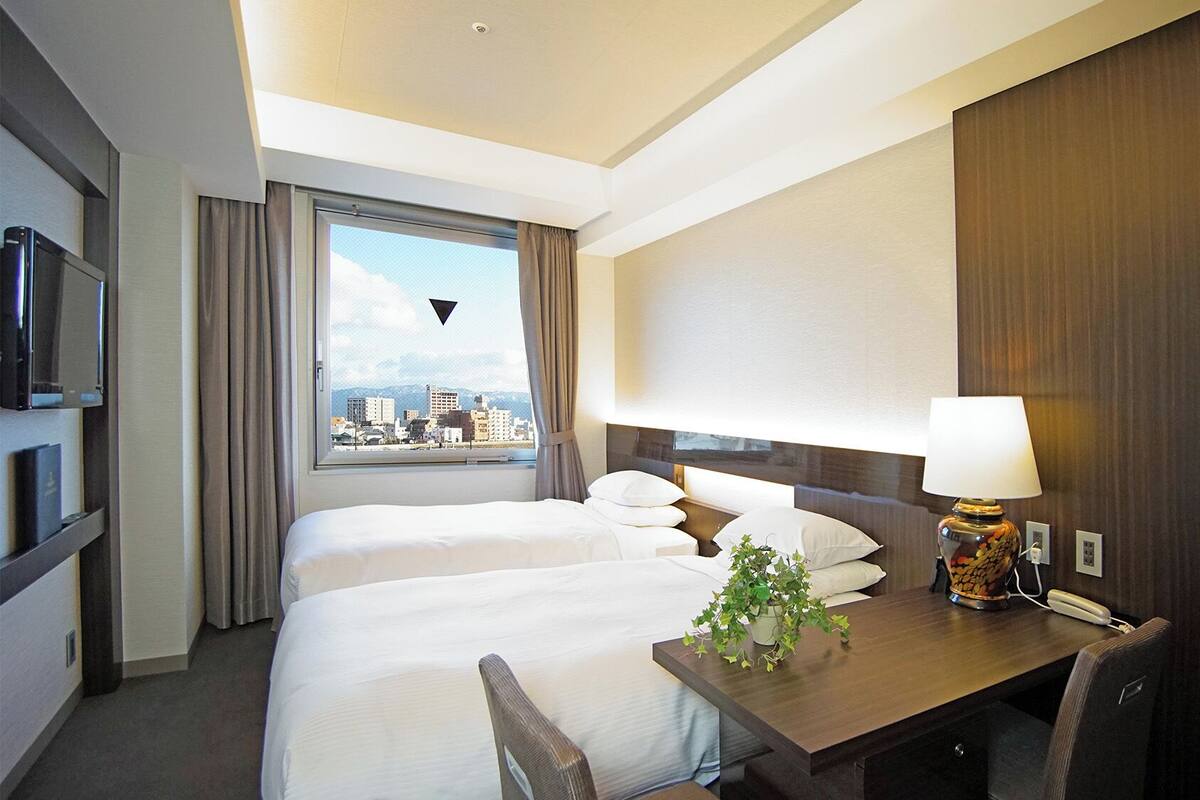 A hotel room with two beds, a desk, a lamp, a TV, and a window with a city view.