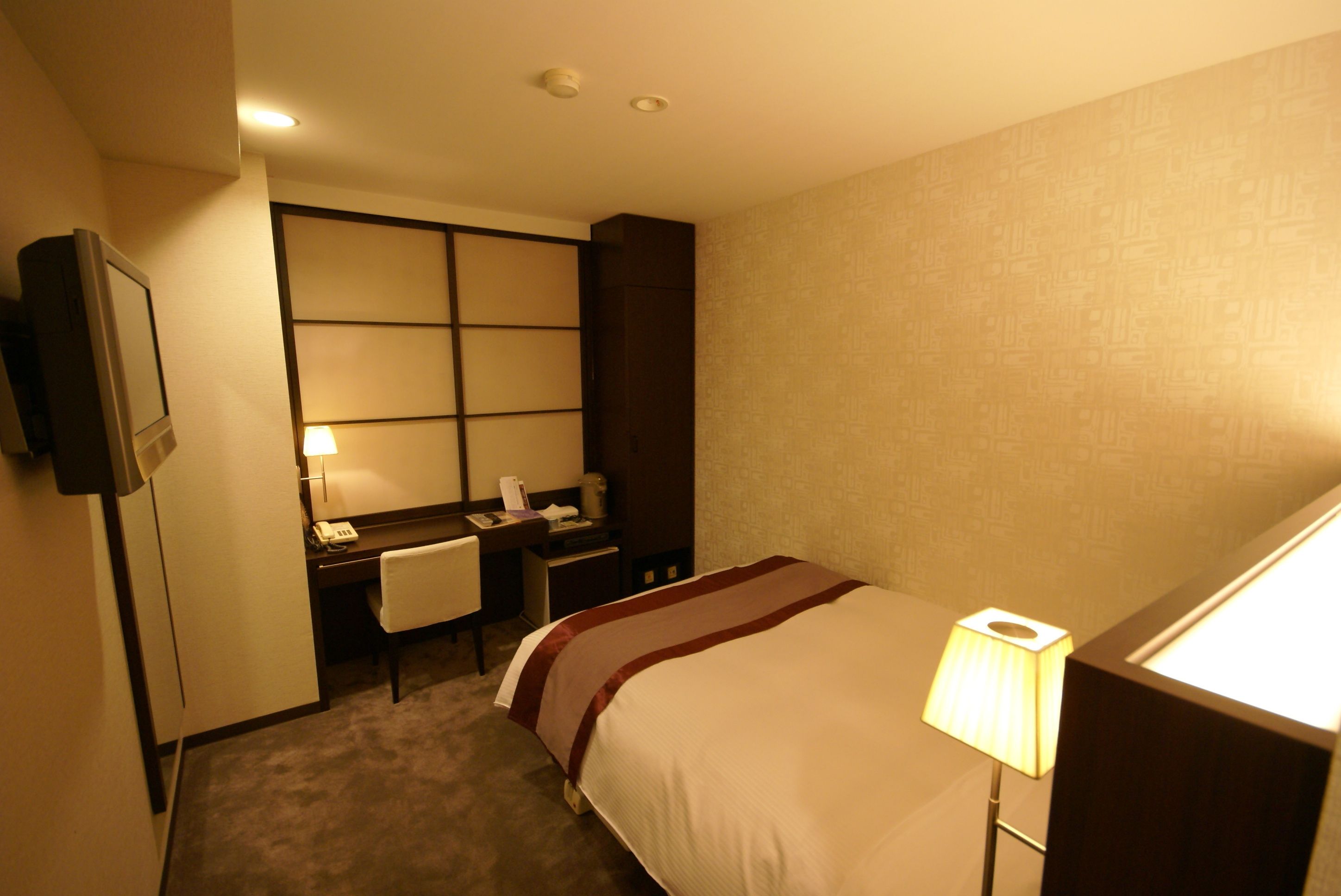 A hotel room with a bed, desk, chair, and a TV on the wall.