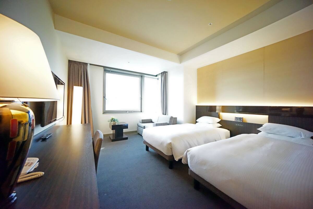 A hotel room with two beds, a desk with a lamp, a chair, a small table with a plant, and a window with curtains.