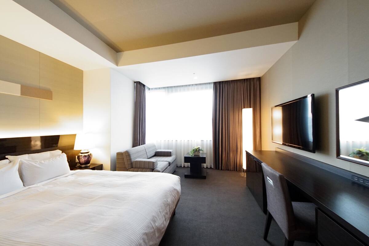A hotel room with a large bed, a desk, a chair, a TV, and a window with curtains.