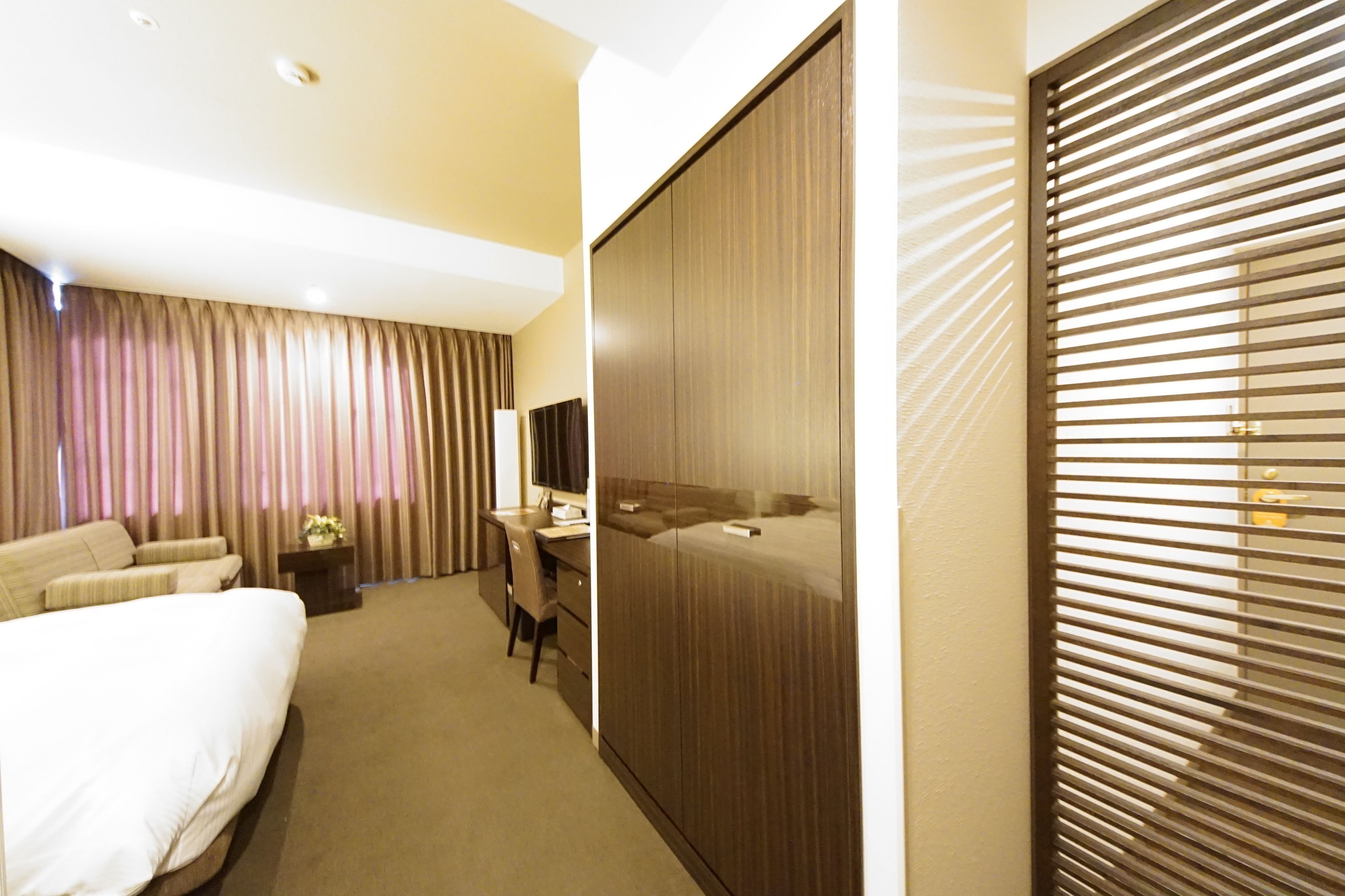 A hotel room with a bed, a desk, a chair, a wardrobe, and a window with blinds.